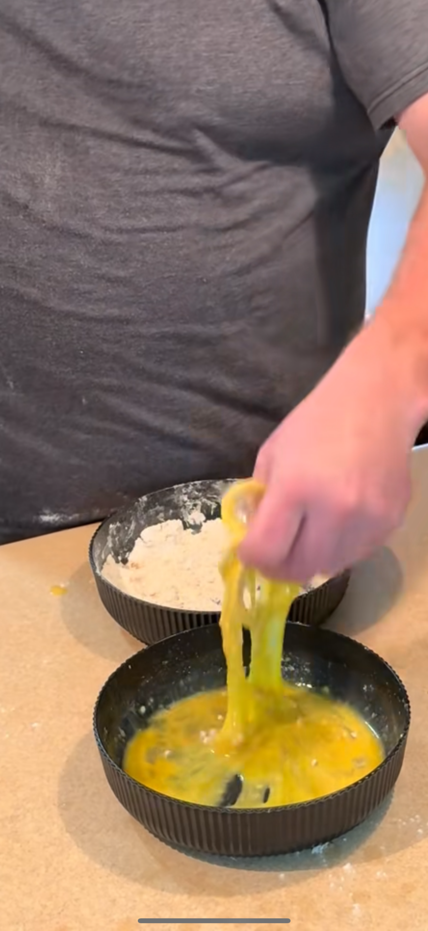 A person dips food into a bowl of beaten eggs with their hand. Another bowl with flour is on the counter nearby. The person is wearing a gray shirt.