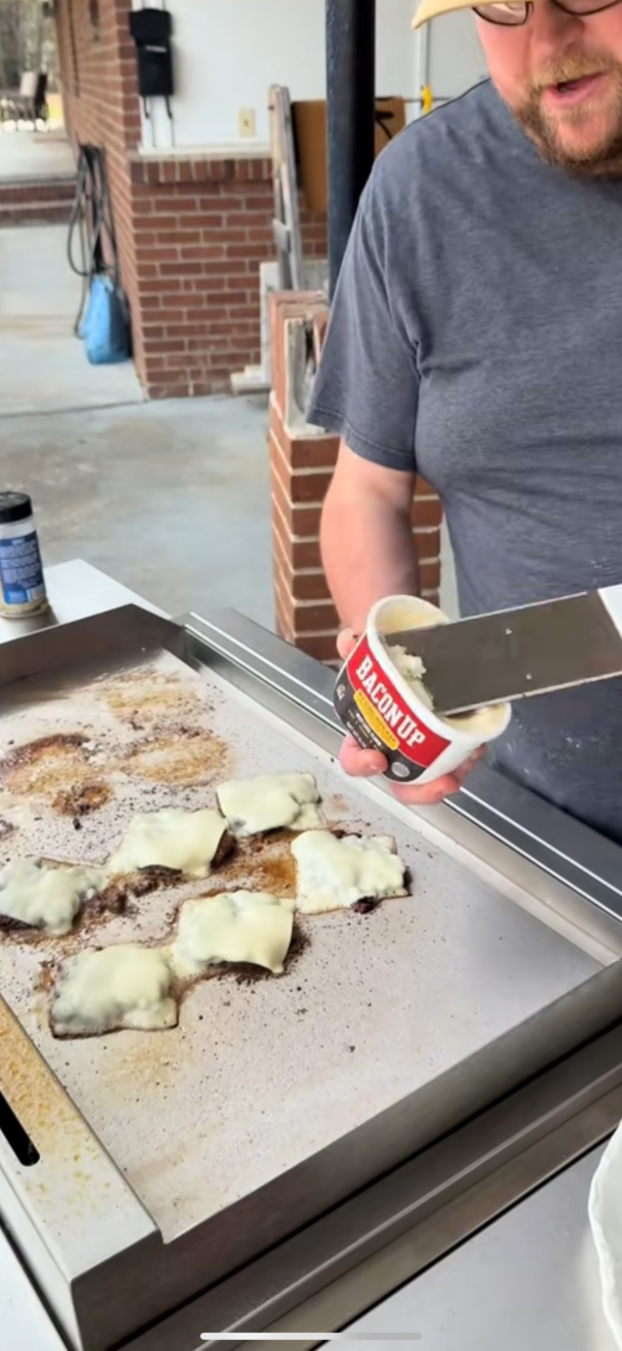 A person wearing glasses and a gray shirt spreads Bacon Up bacon grease onto a flat top grill with cheeseburgers cooking, each topped with melted cheese.