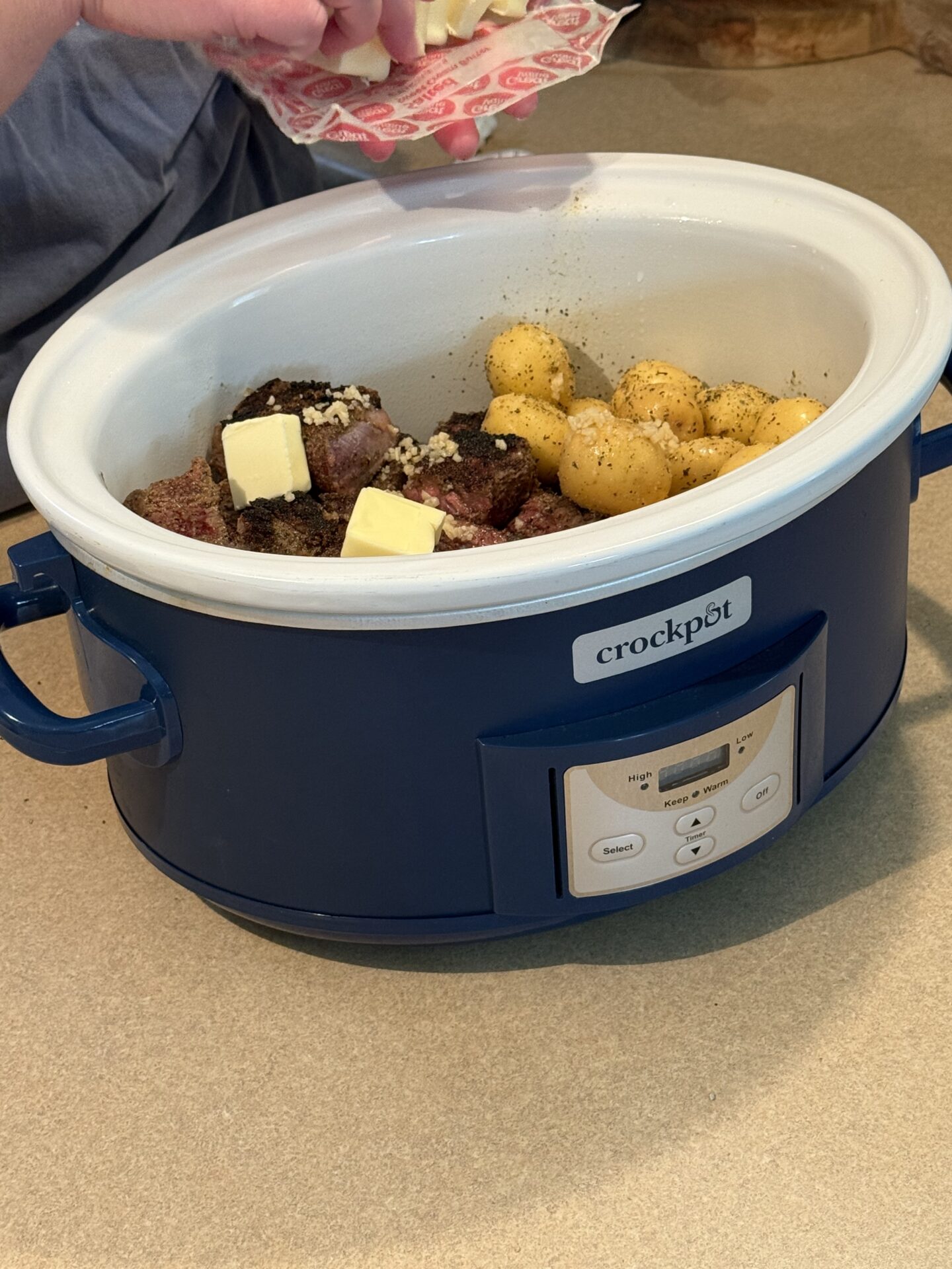 A hand adds cubes of butter to a blue Crockpot filled with seasoned meat, small yellow potatoes, and minced garlic, all set on a kitchen countertop.