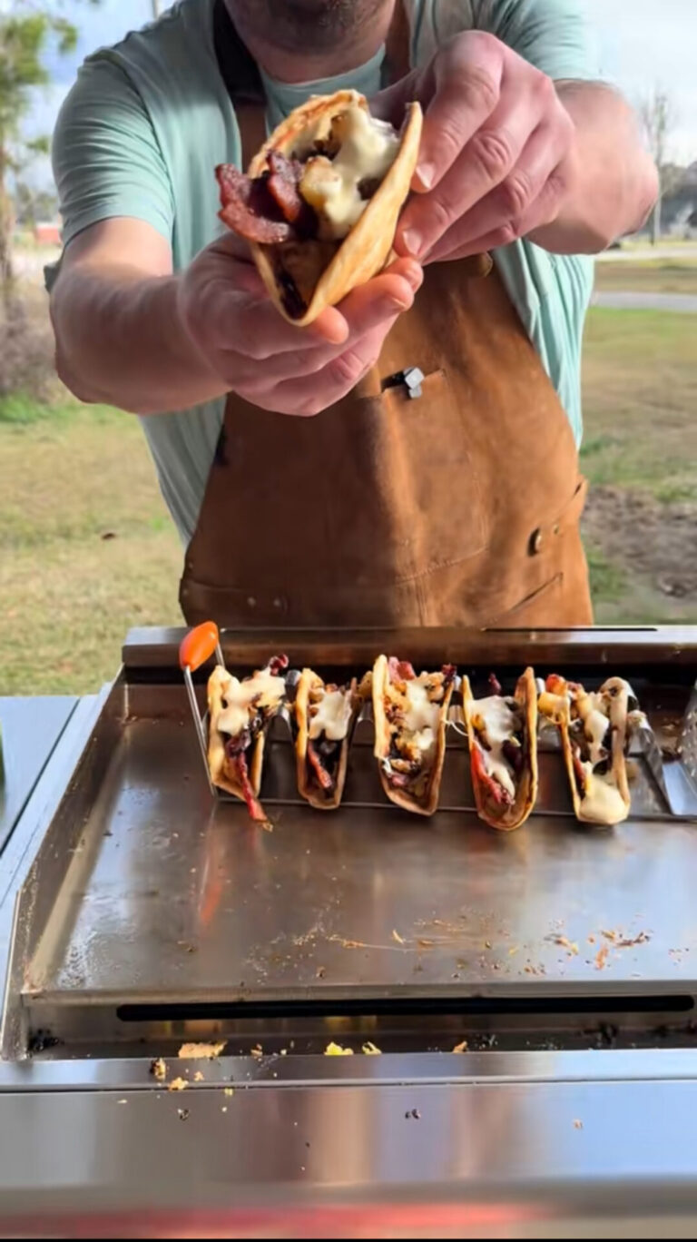 A person wearing a brown apron holds a cheesy bacon taco toward the camera, with several similar tacos lined up on a grill outdoors.