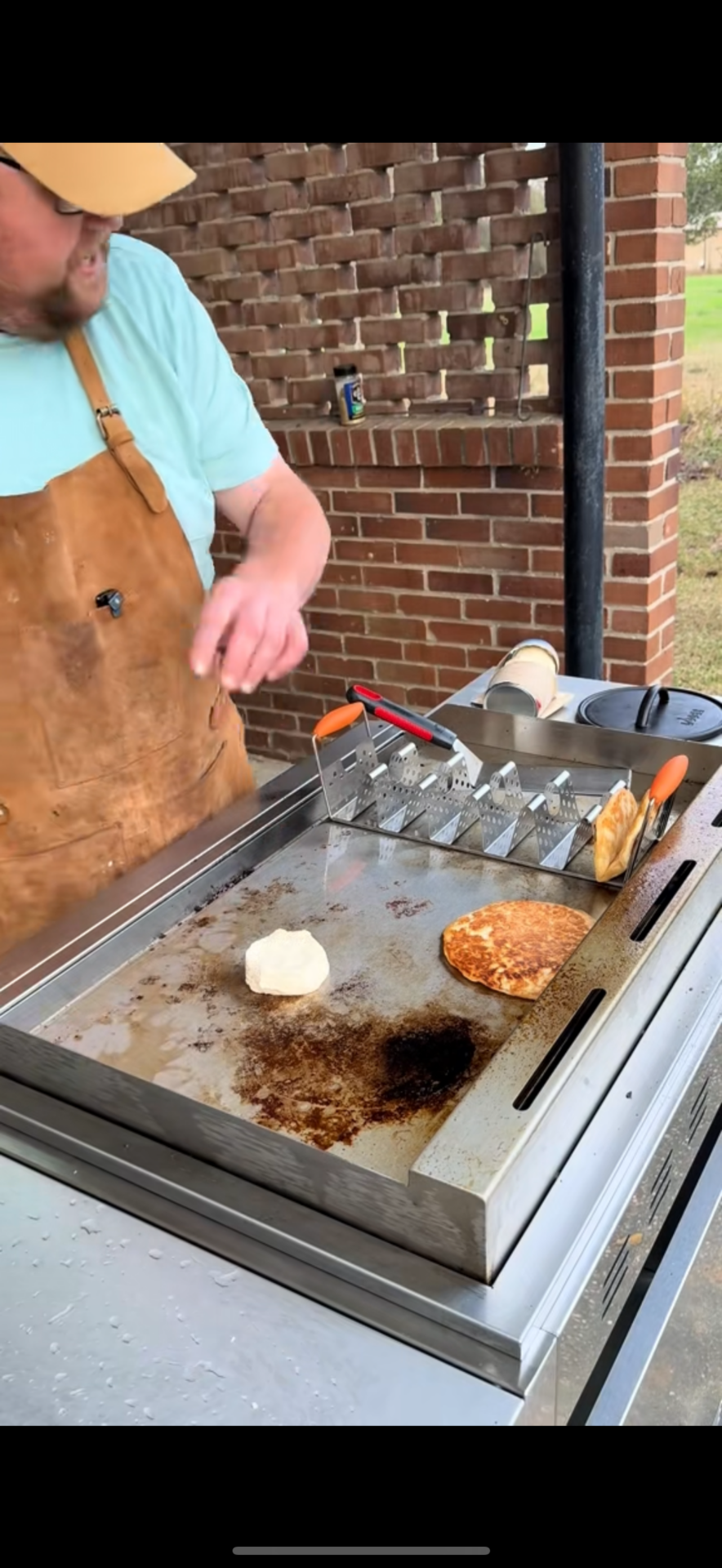 A person wearing a tan apron and light blue shirt cooks food on a flat-top grill outdoors. There is a burger patty, a dough ball, and bread on the grill, with utensils and a brick wall in the background.