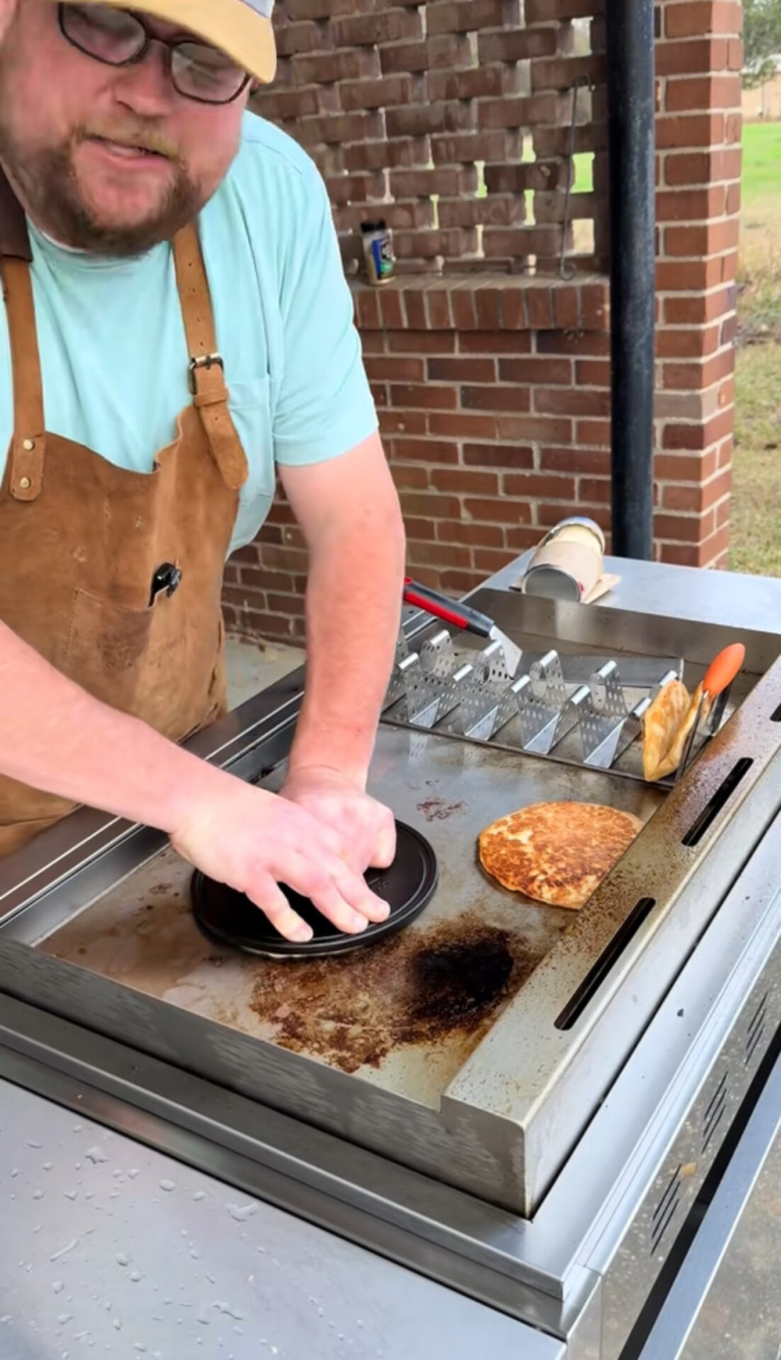 A person in a brown apron and blue shirt presses a burger patty with a metal press on a griddle outdoors. There is a cooked patty, a spatula, and a brick wall in the background.