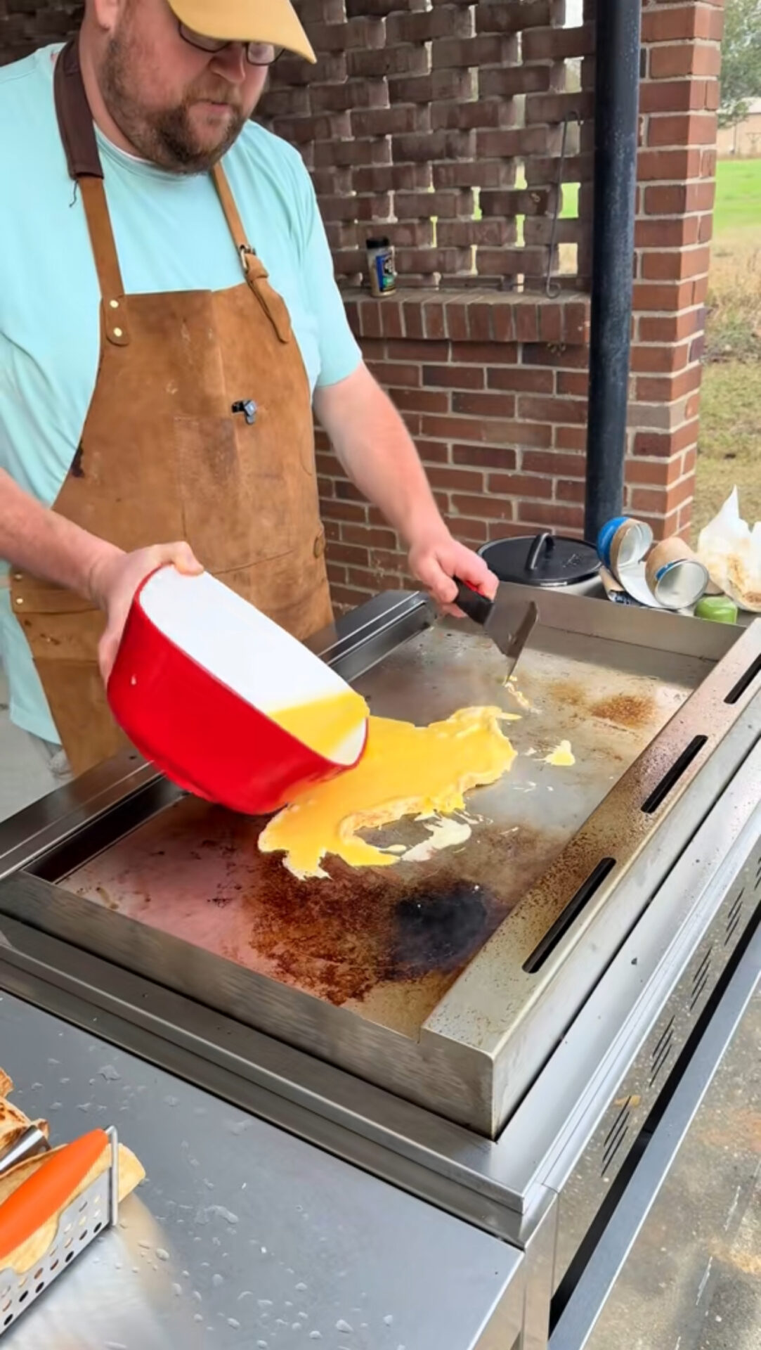 A man in a brown apron pours beaten eggs from a red bowl onto a flat-top outdoor griddle, preparing to cook scrambled eggs. Brick walls and greenery are visible in the background.