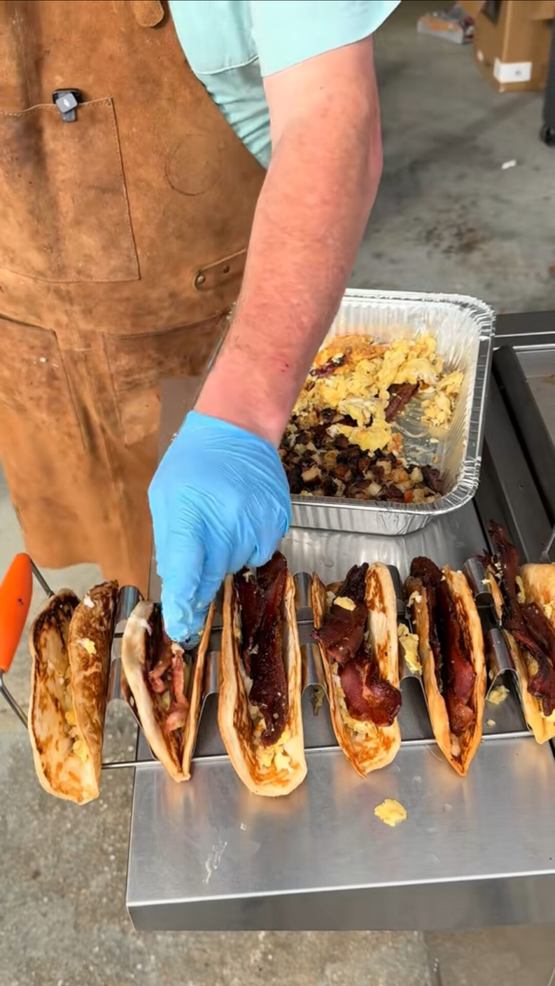 A person wearing a blue glove and a brown apron fills tacos with bacon, standing beside a tray of scrambled eggs and potatoes on a stainless steel counter.