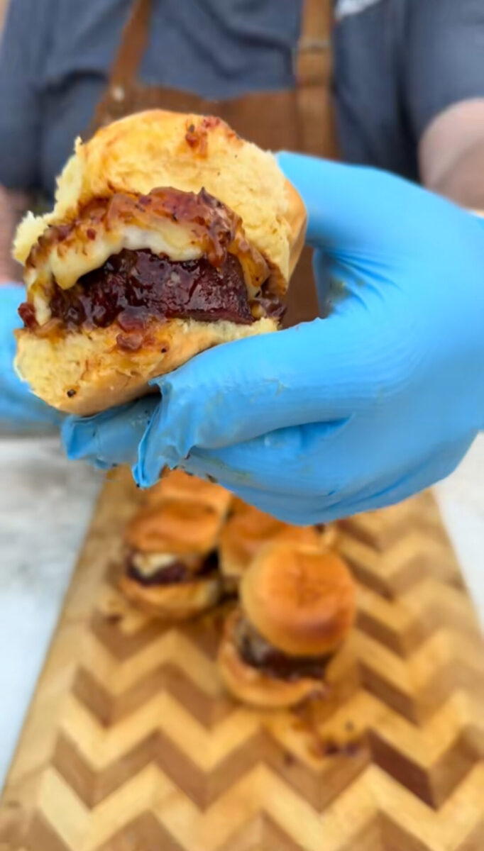 A person wearing blue gloves holds up a small burger with cheese and sauce. More sliders are arranged on a zigzag-patterned wooden board in the background.