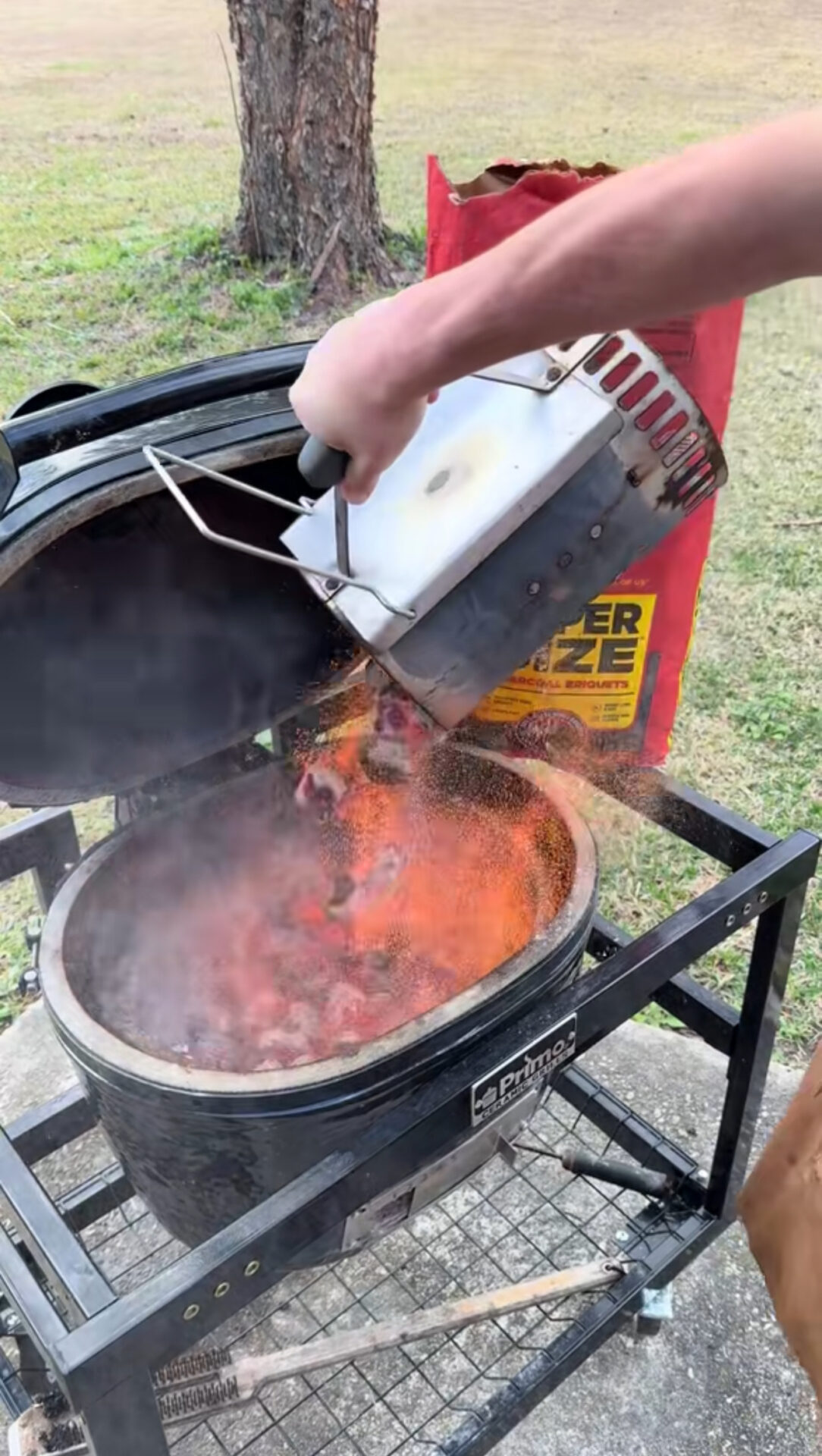 A person pours hot charcoal from a metal chimney starter into a ceramic grill outdoors, with flames and smoke rising; a bag of charcoal sits nearby on the grass.