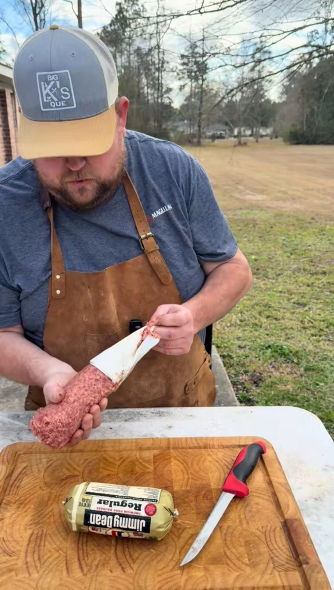 A man in a brown apron and blue shirt prepares ground meat outdoors, peeling the wrapper from a tube of Jimmy Dean sausage on a cutting board. A knife with a red handle is nearby. Trees and grass are in the background.