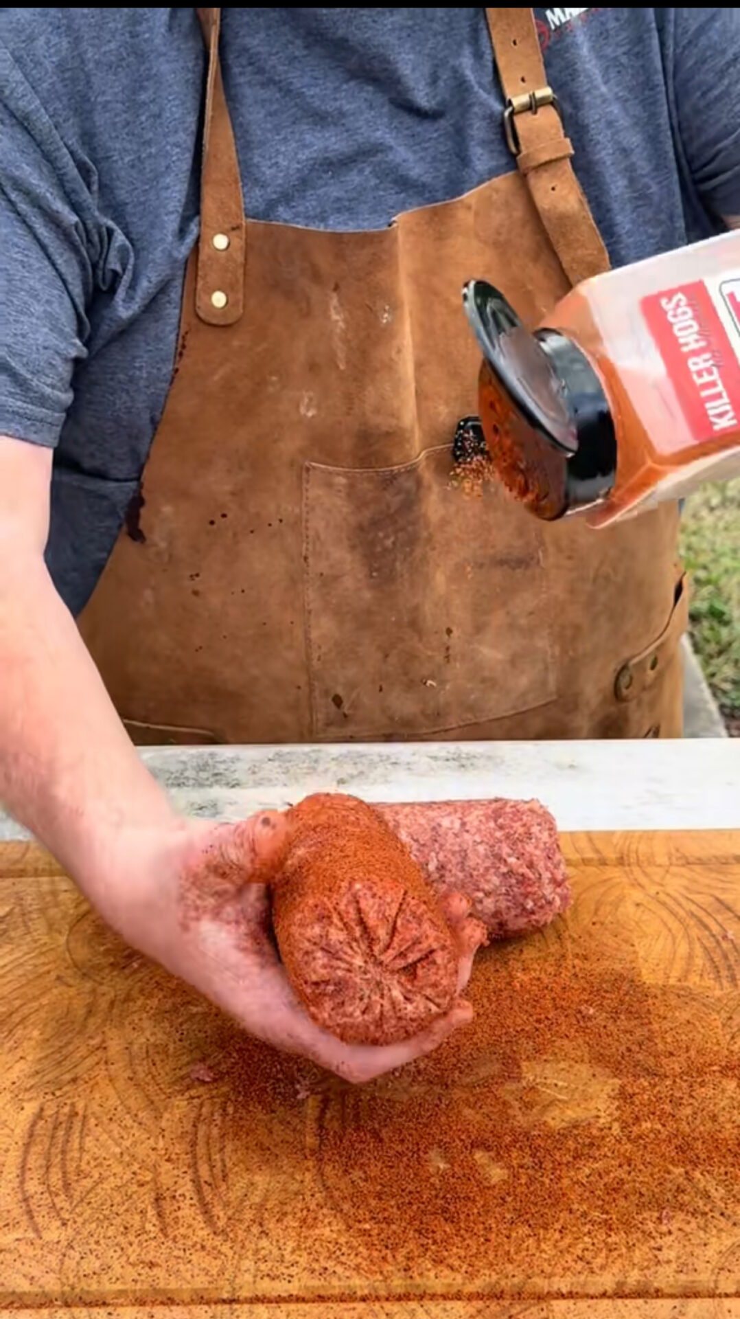 A person wearing a brown apron sprinkles seasoning labeled Killer Hogs onto a raw sausage held over a wooden cutting board.