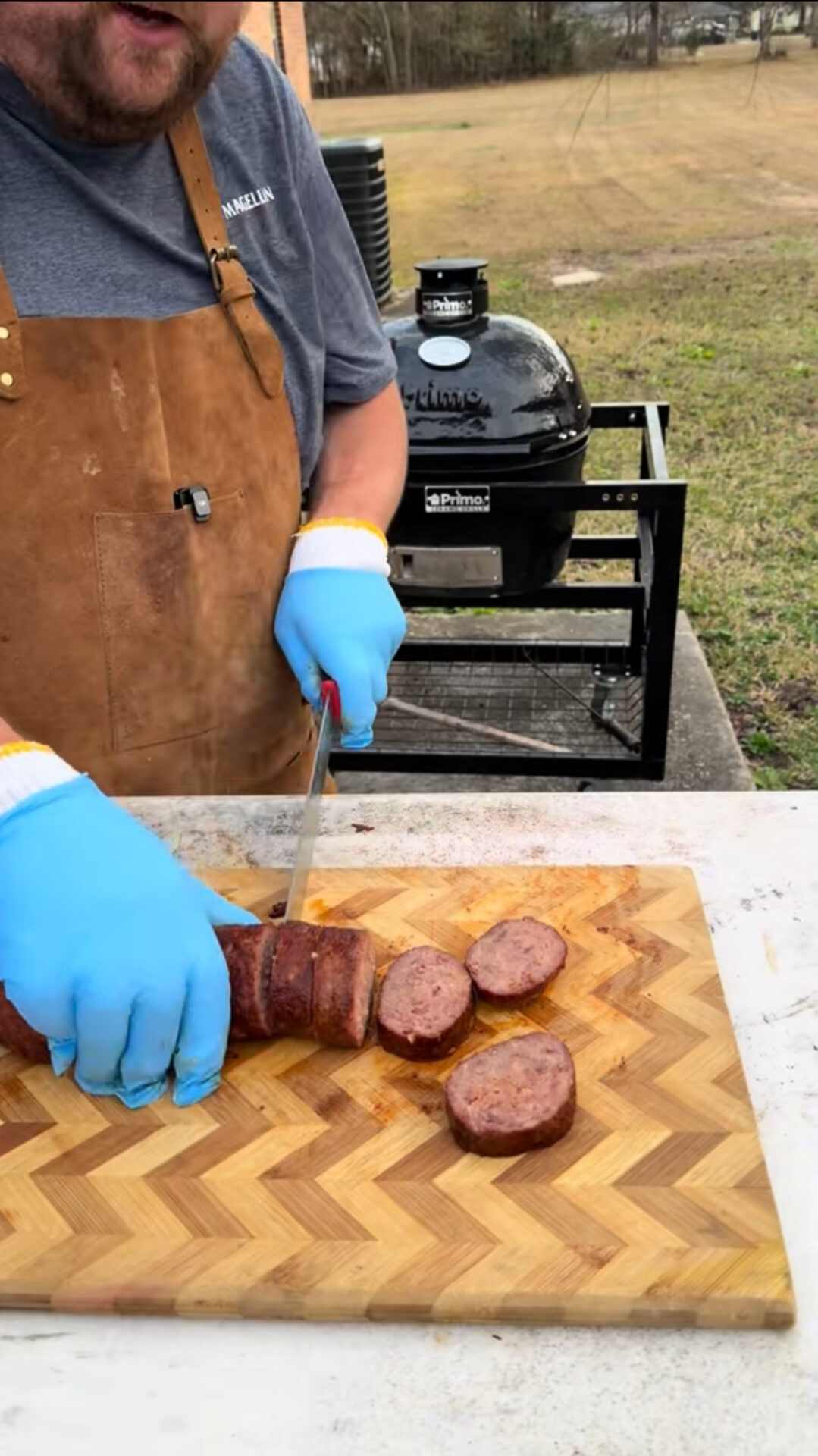 A person wearing blue gloves and a brown apron slices smoked sausage on a wooden cutting board outdoors, with a black grill and grassy area in the background.
