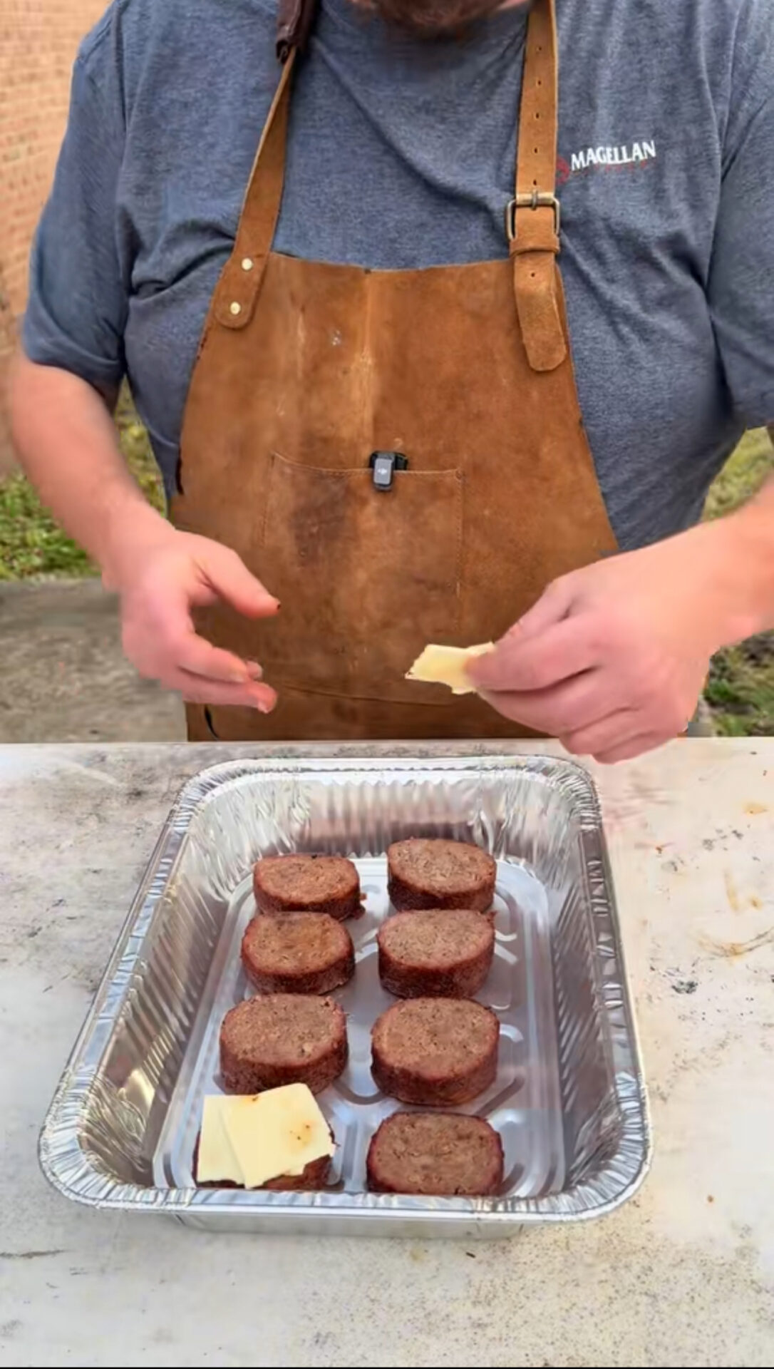 A person wearing a brown apron and gray shirt places slices of cheese on top of round pieces of cooked meat arranged in a foil tray.