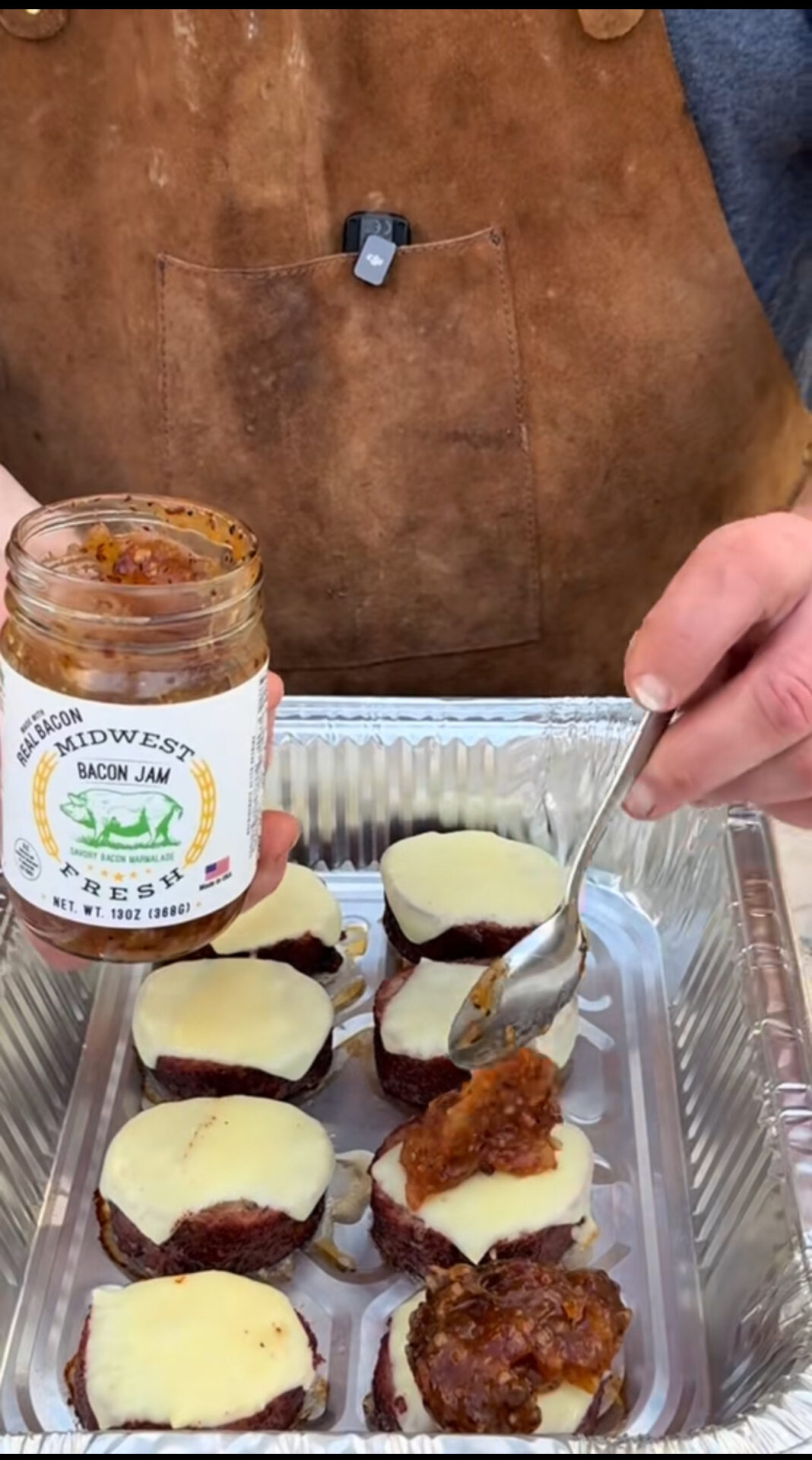 A person wearing a brown apron spreads bacon jam from a jar onto cheese-topped meat rounds in a foil tray, preparing a savory dish.
