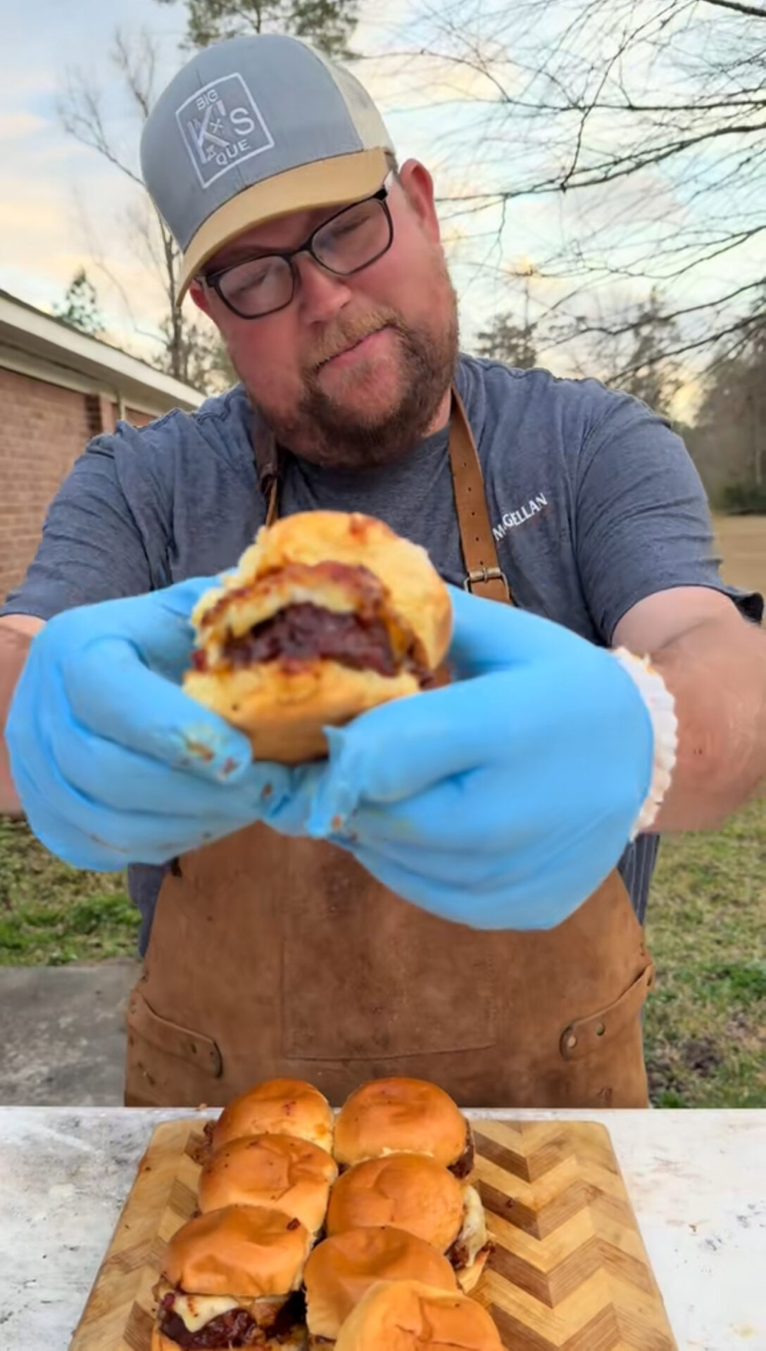A man wearing a cap, glasses, blue gloves, and an apron holds a cheeseburger slider towards the camera with more sliders on a wooden board in front of him, outdoors.