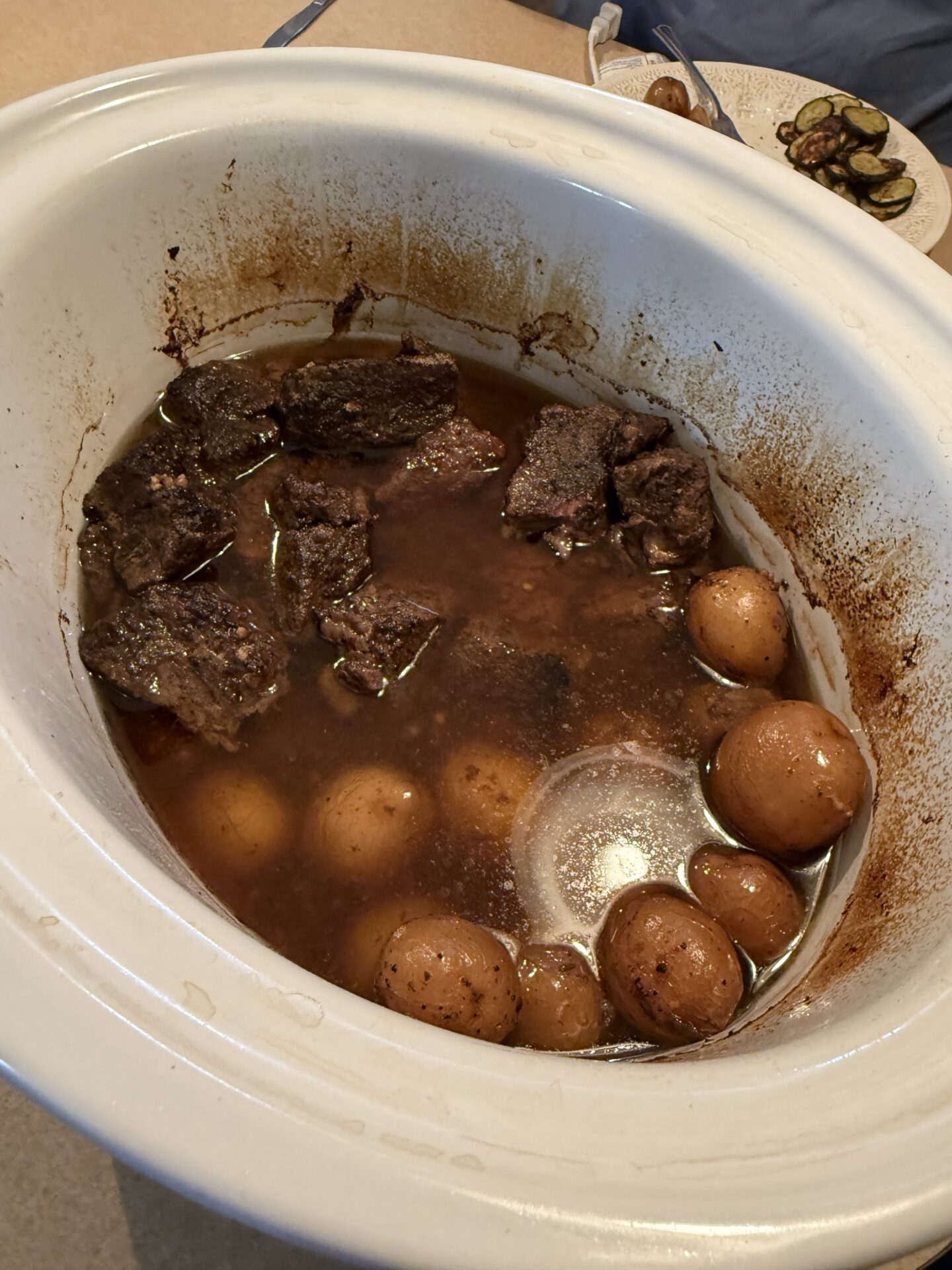 A white slow cooker filled with cooked beef chunks, whole baby potatoes, and brown broth. The food appears well-cooked, and the sides of the cooker are stained with sauce. A plate of sliced vegetables is in the background.
