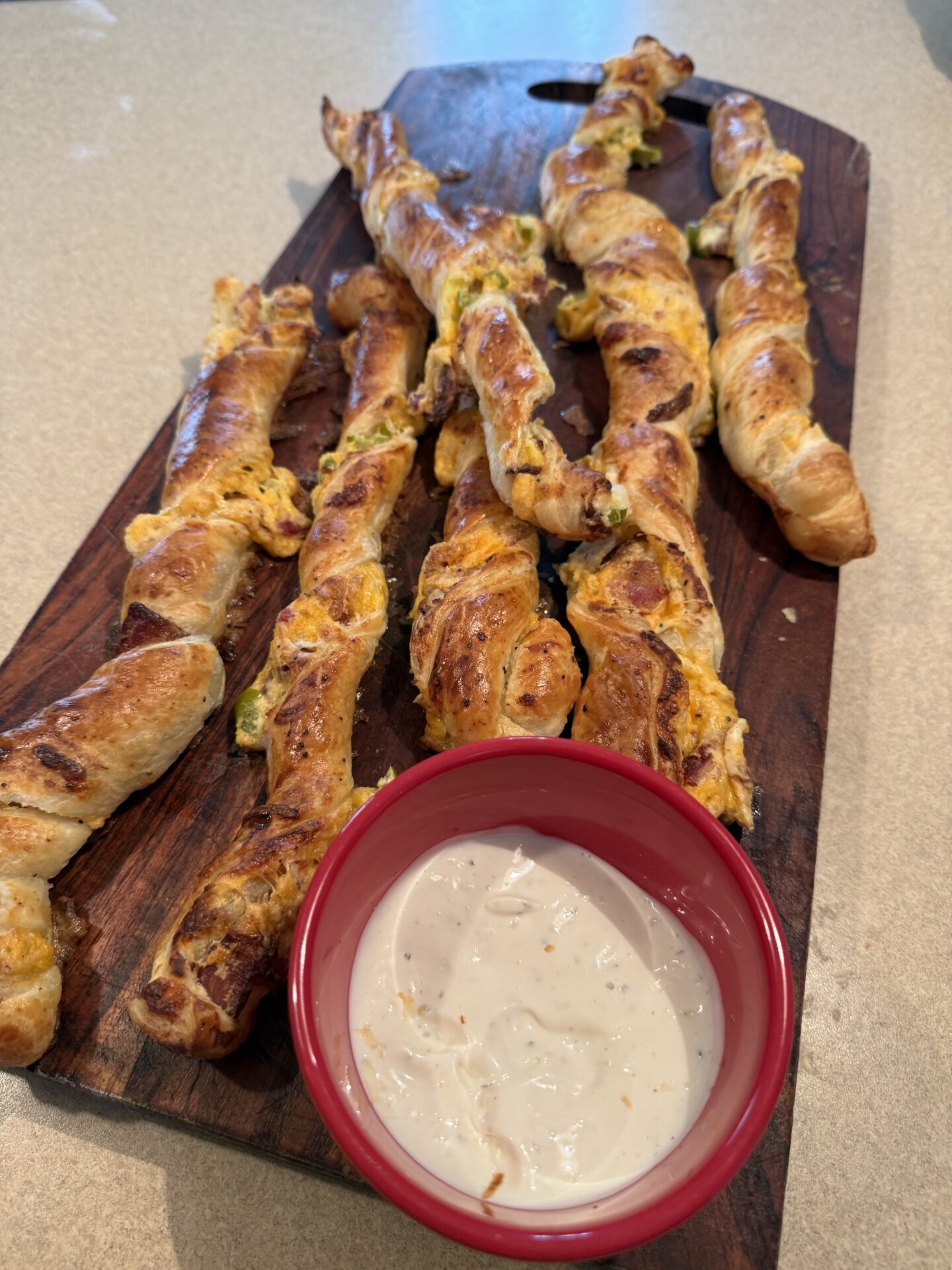 Six twisted cheese pastry sticks are arranged on a wooden board with a small red bowl of creamy dipping sauce in front. The pastries appear golden brown and are served on a light countertop.