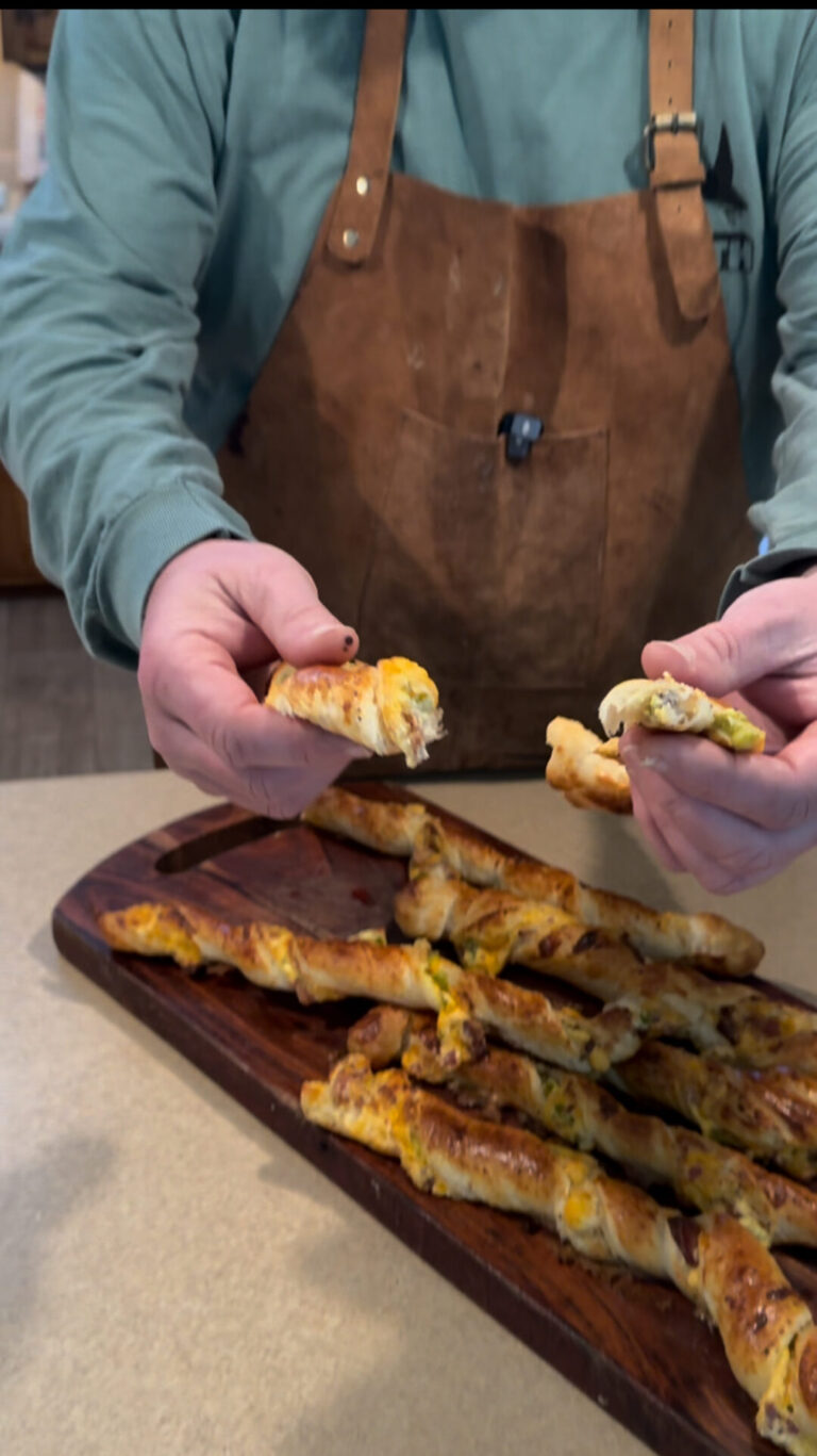 A person wearing a brown apron and green shirt holds a split cheesy breadstick, with several more breadsticks on a wooden cutting board on the counter in front of them.