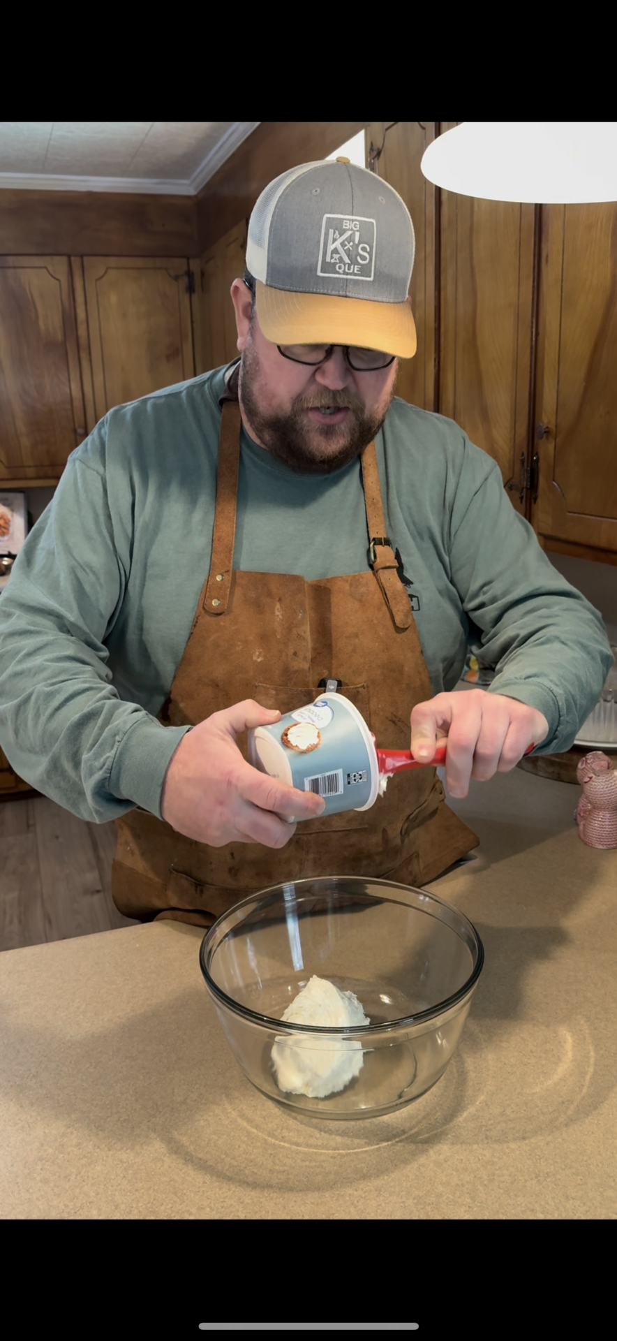 A bearded man in glasses and a brown apron scoops sour cream from a container into a glass bowl on a kitchen counter, with wooden cabinets in the background.