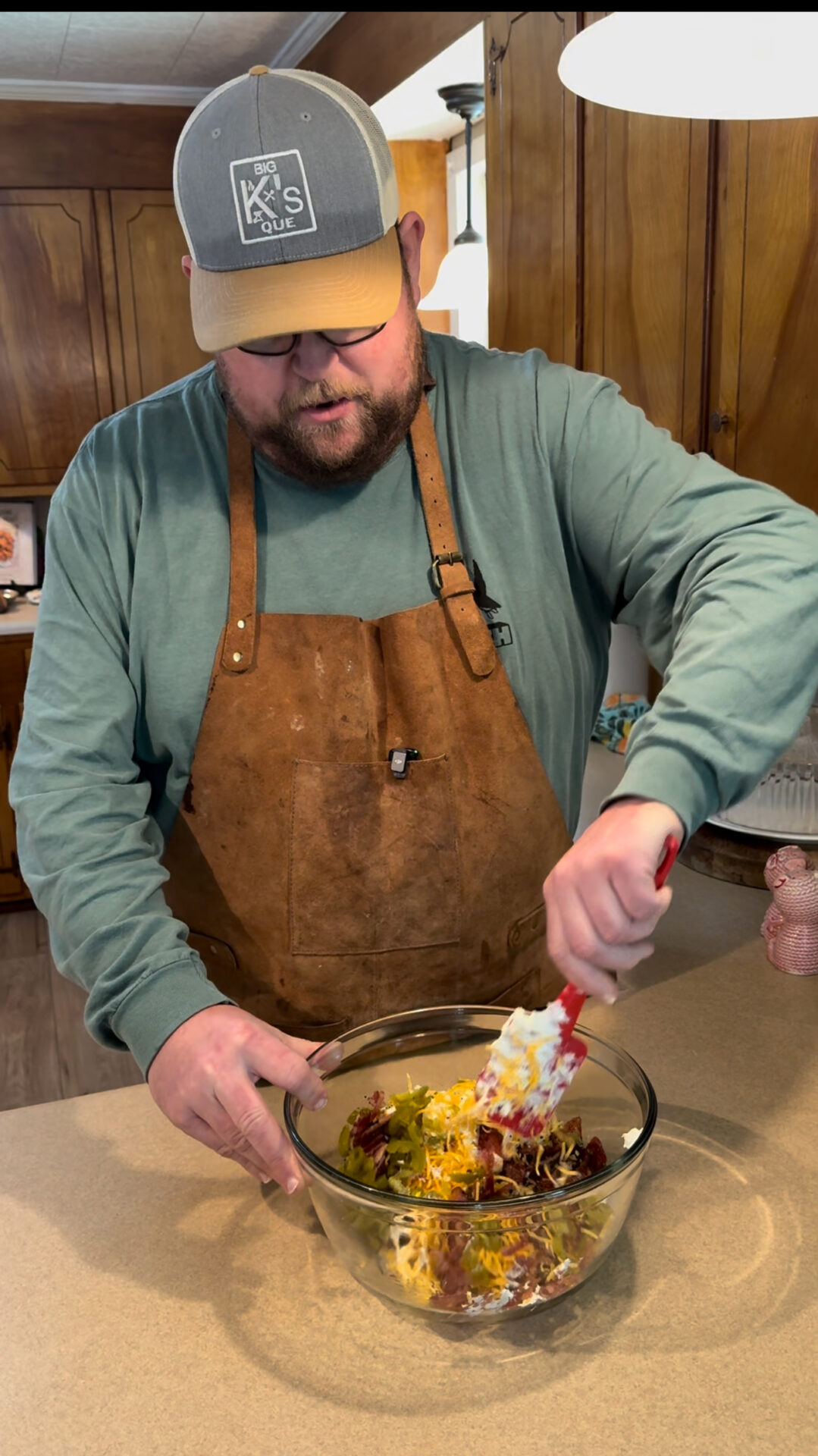 A man wearing glasses, a green shirt, brown apron, and gray cap is mixing ingredients in a glass bowl on a kitchen counter. He uses a spatula to combine shredded cheese and other items into jalapeno popper twists. Wooden cabinets are visible in the background.