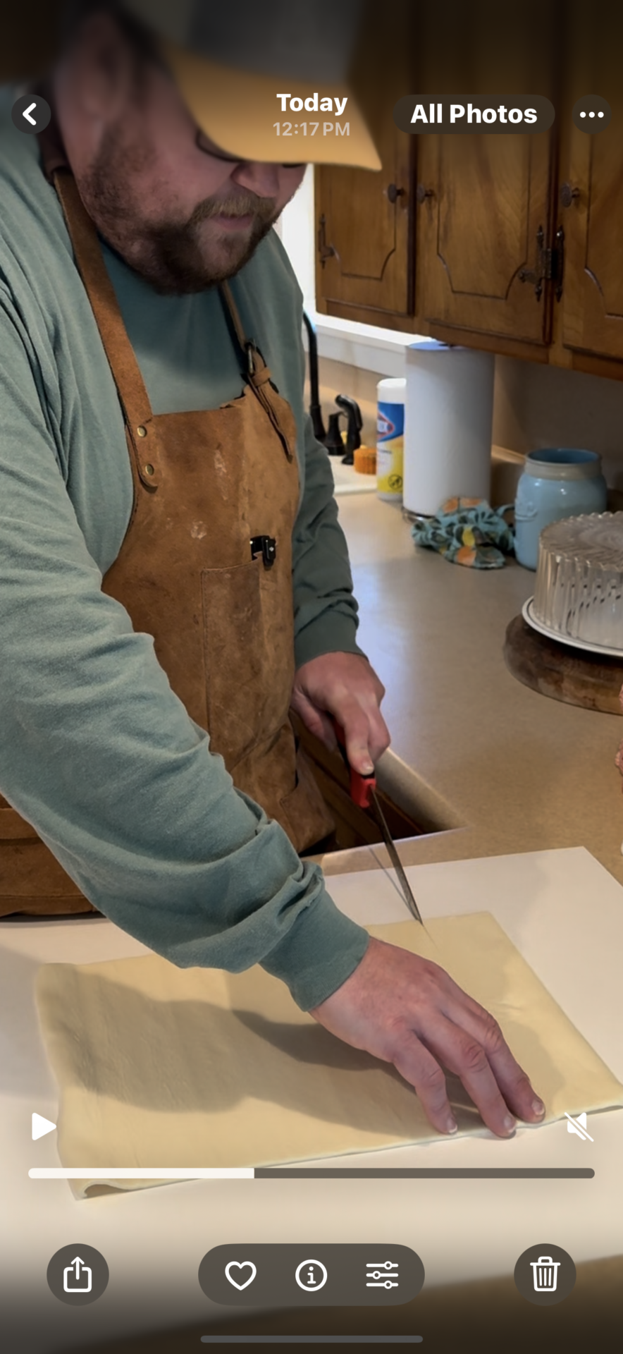 A person wearing a brown apron and green shirt cuts a sheet of dough on a kitchen counter with a red-handled knife. Wooden cabinets, a cake, and kitchen items are visible in the background.