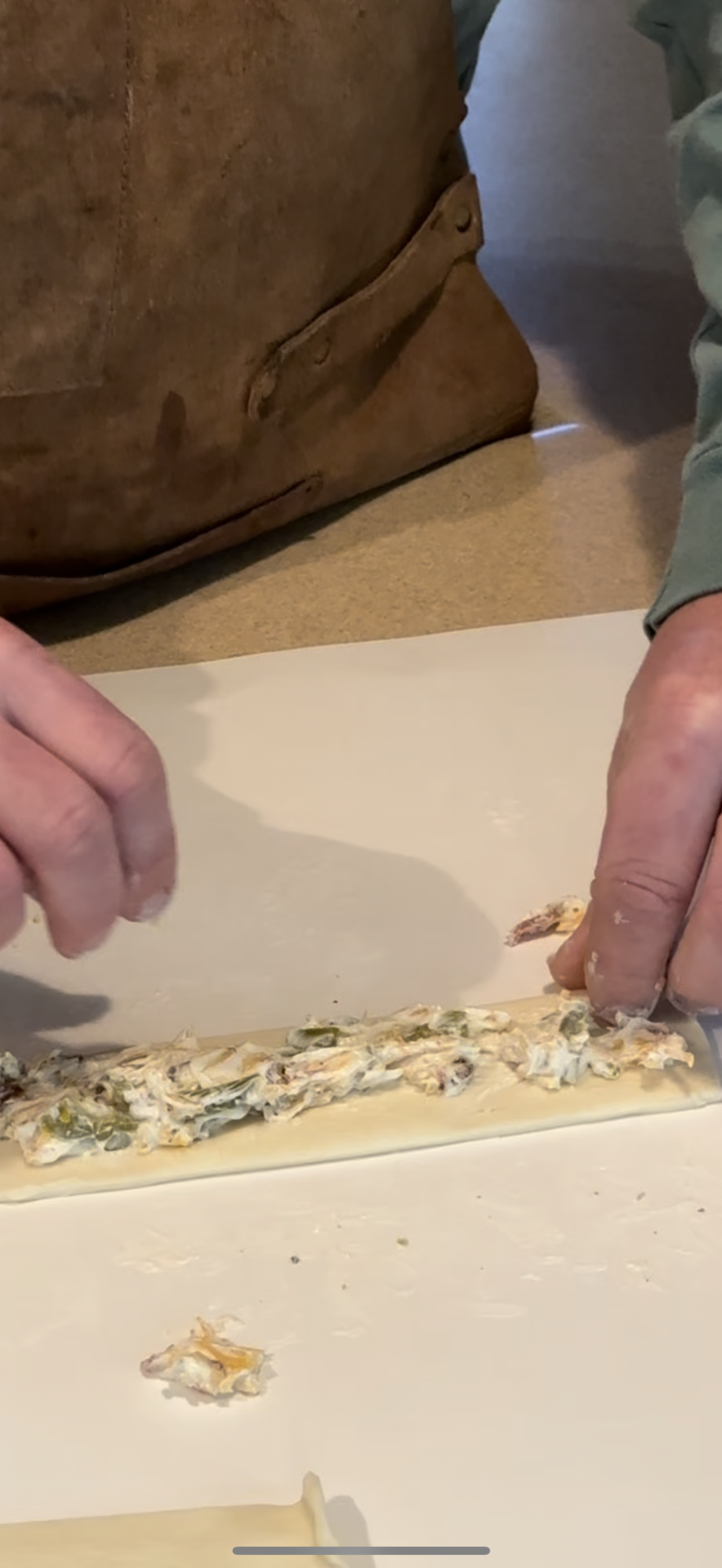A person in a brown apron is rolling a sheet of dough filled with a creamy mixture on a white surface, preparing to make a rolled pasta or pastry dish.