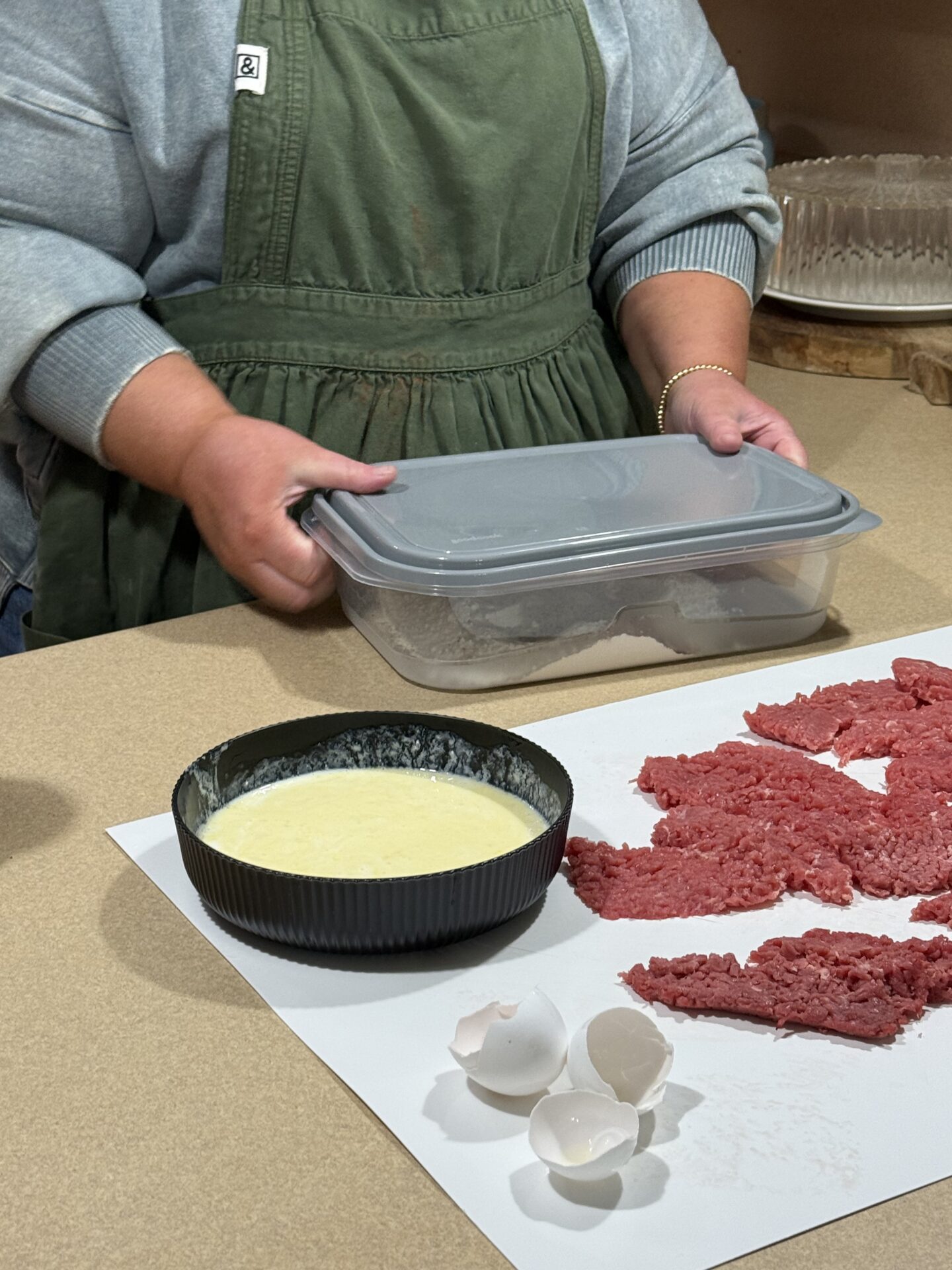 A person in a green apron holds a plastic container next to a countertop with raw ground beef, a black bowl of beaten eggs, and cracked eggshells on a white surface.