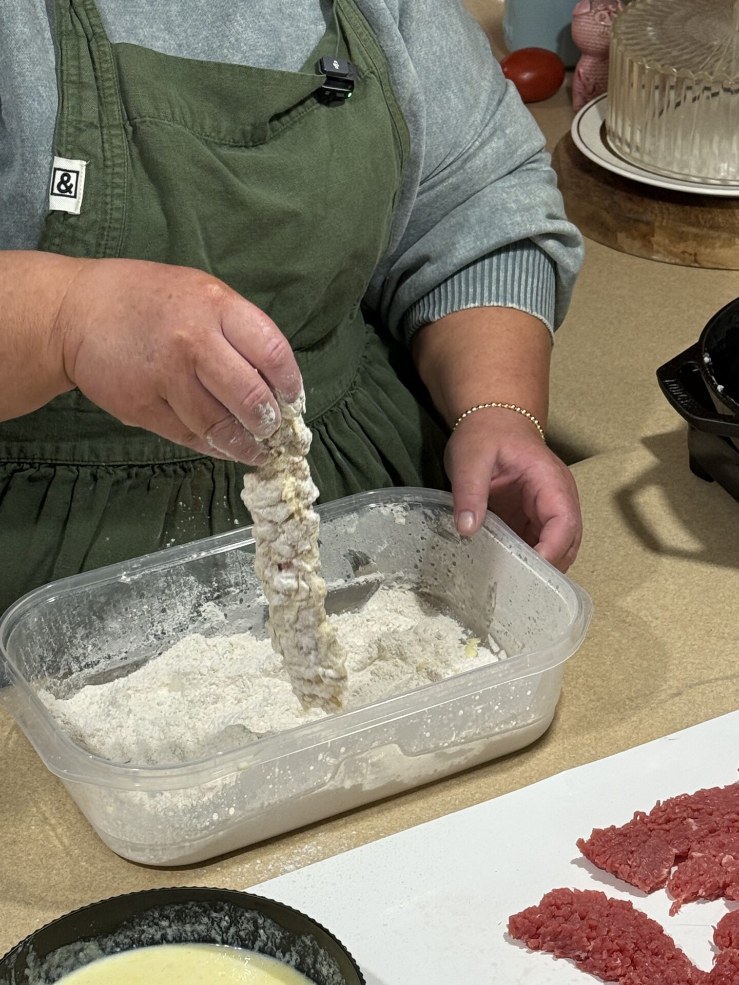 A person in a green apron dips a strip of raw meat into a container of flour, preparing to bread it. Nearby, there are more strips of raw meat and a bowl with a yellow liquid, likely egg wash.