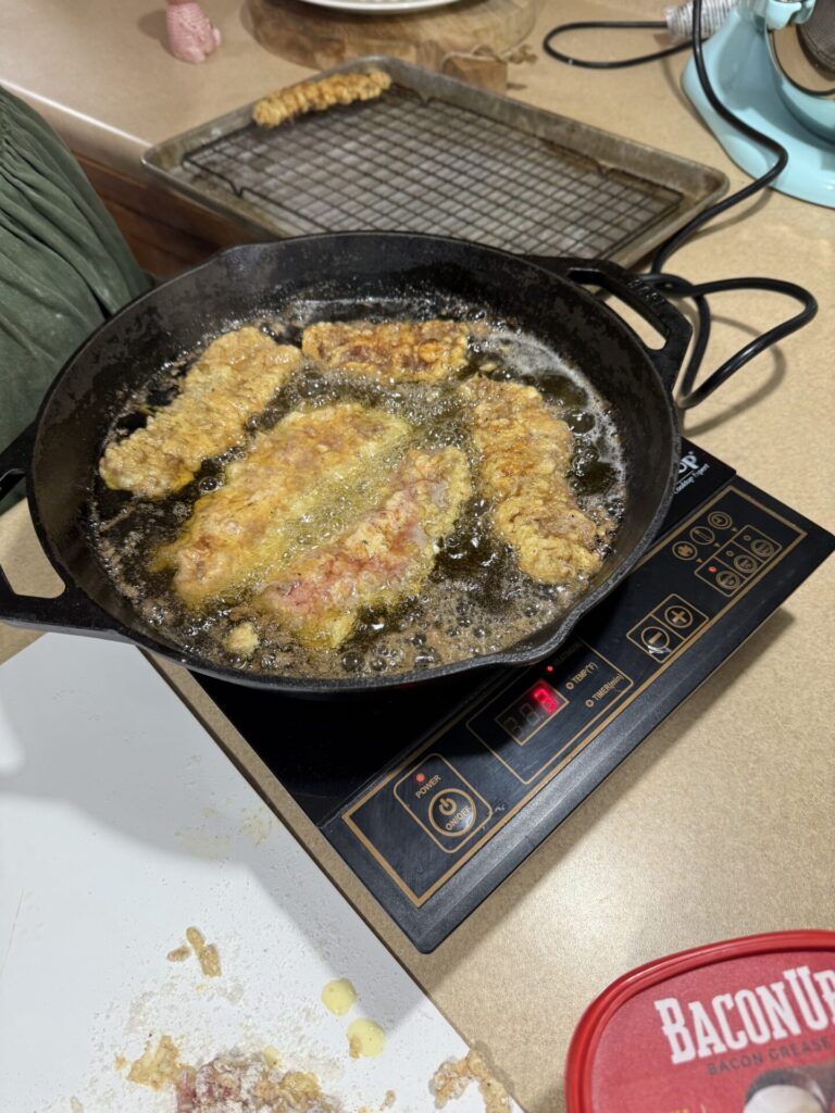 Chicken strips being deep-fried in a cast iron skillet on an induction cooktop, with a cooling rack and other kitchen items visible on the counter nearby.