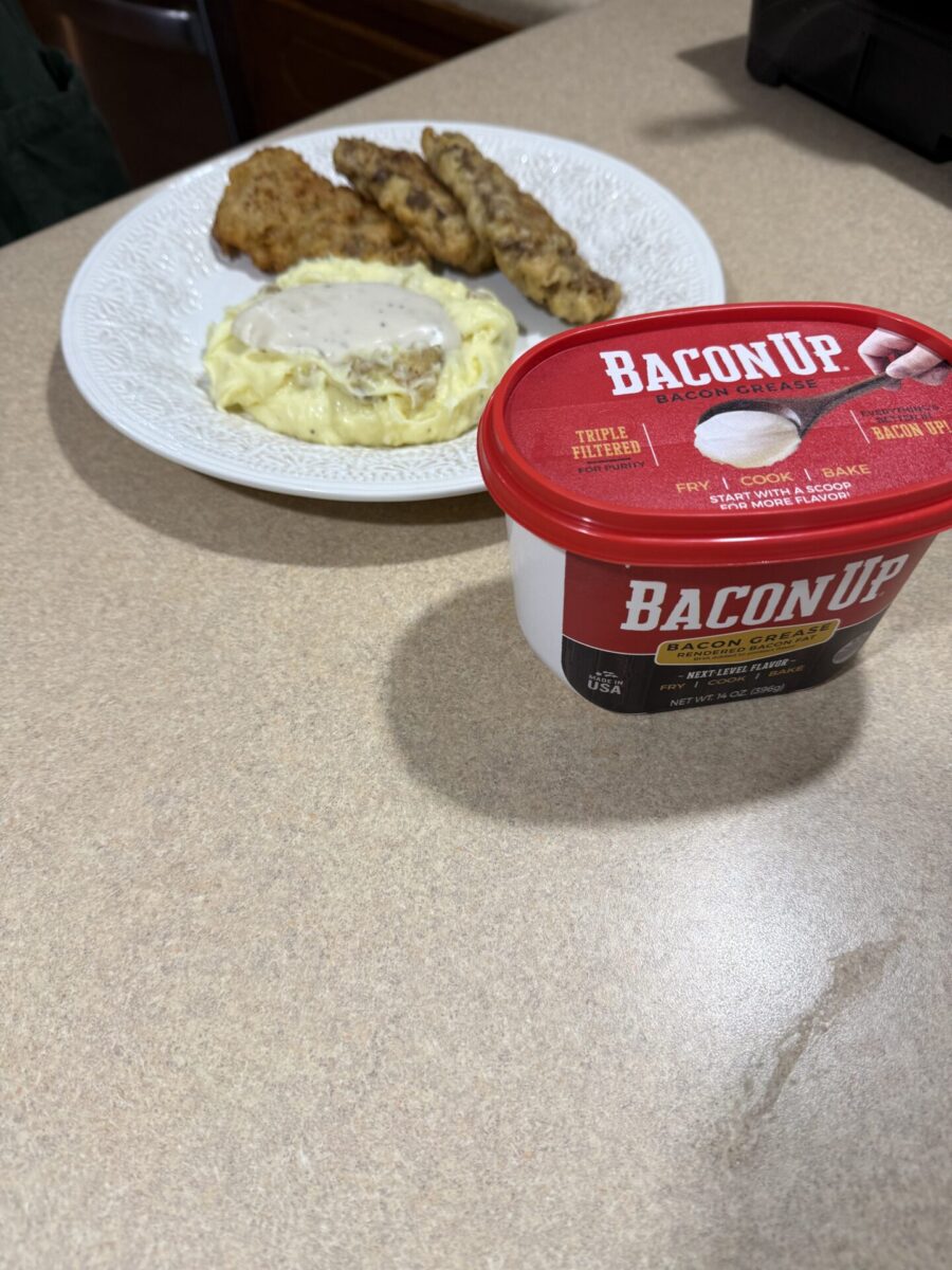 A plate of mashed potatoes with gravy, a breaded patty, and chicken tenders sits on a counter. In the foreground is a container of Bacon Up bacon grease. Recipe is for Country Fried Steak Fingers