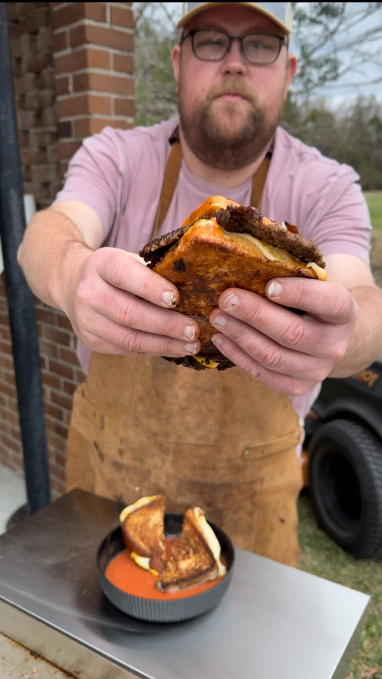 A man wearing glasses, a pink shirt, and a brown apron holds a grilled cheese sandwich toward the camera. On the table in front of him is a bowl of tomato soup with another grilled cheese sandwich.