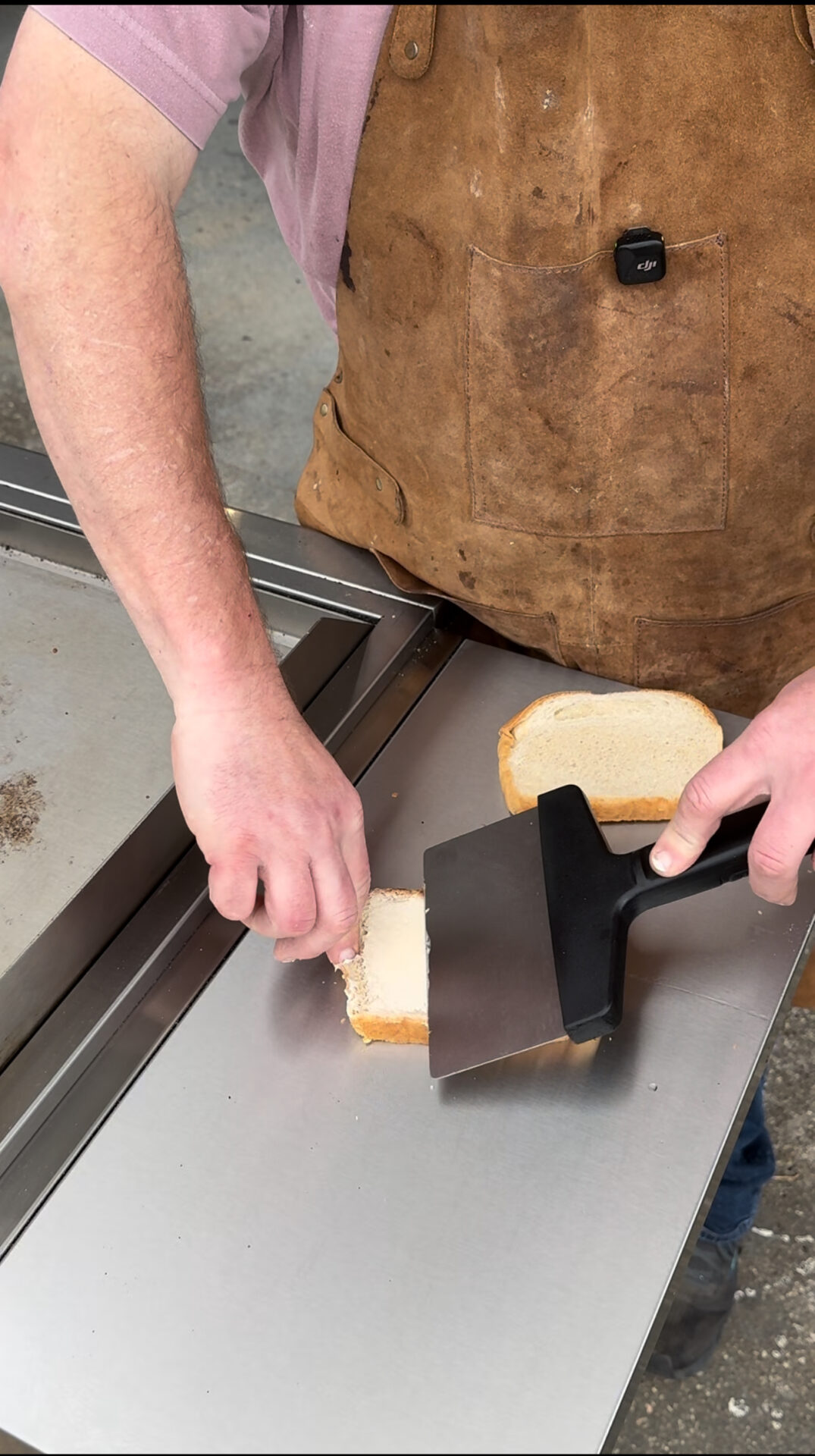 A person wearing a brown apron slices a piece of white bread with a spatula on a stainless steel surface near a griddle.