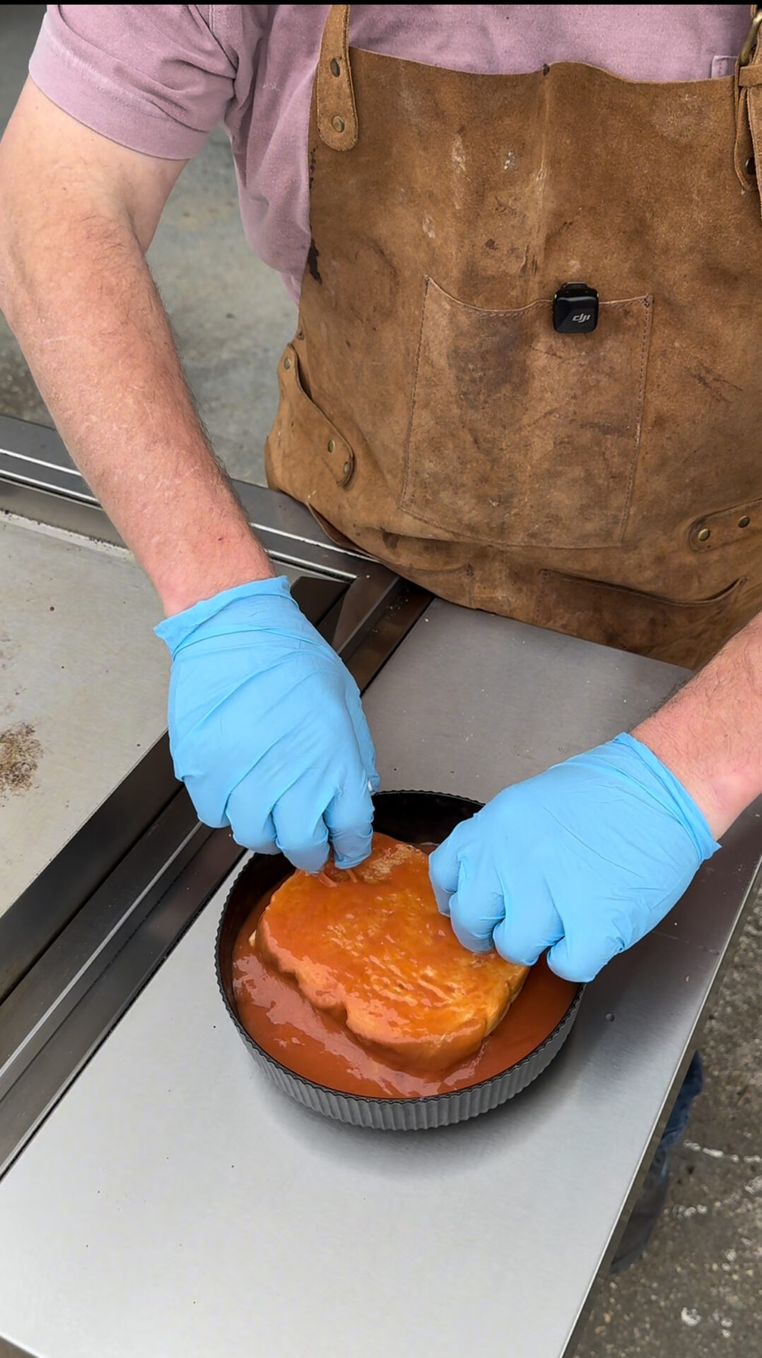 A person wearing blue gloves and a brown apron dips a piece of raw meat into a round pan filled with orange marinade on a metal surface.