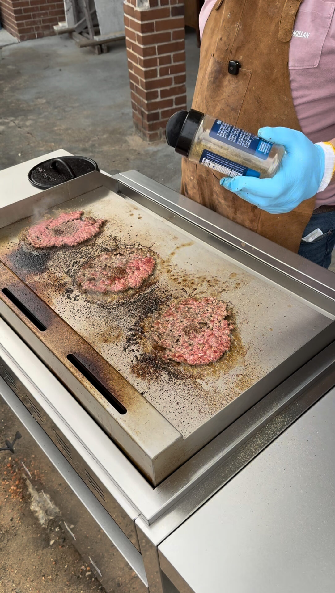 A person wearing blue gloves and a brown apron is holding a seasoning bottle while cooking three burger patties on a flat-top grill. The patties are sizzling and partially cooked.
