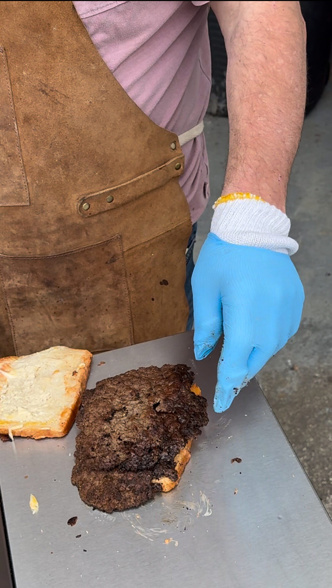 A person wearing a brown apron, blue glove, and a protective wristband prepares a sandwich with a slice of bread and a large, well-cooked meat patty on a metal surface.