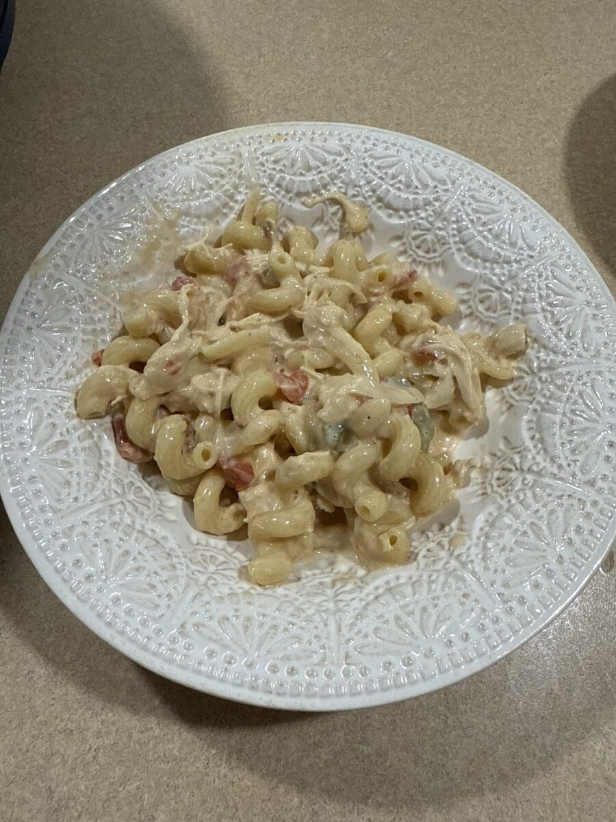 A white plate with an intricate design holds a serving of creamy macaroni pasta mixed with shredded chicken and diced tomatoes on a beige countertop.