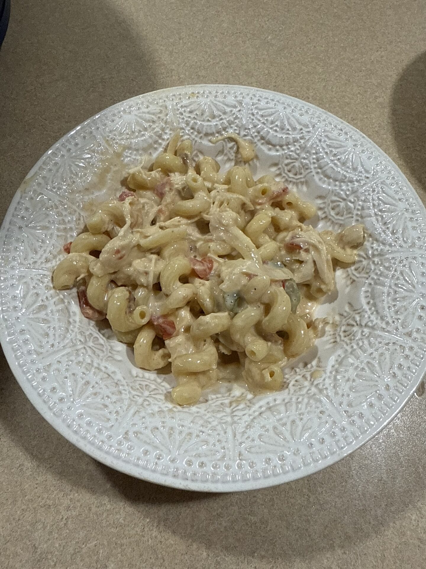 A white plate with an intricate design holds a serving of creamy macaroni pasta mixed with shredded chicken and diced tomatoes on a beige countertop.