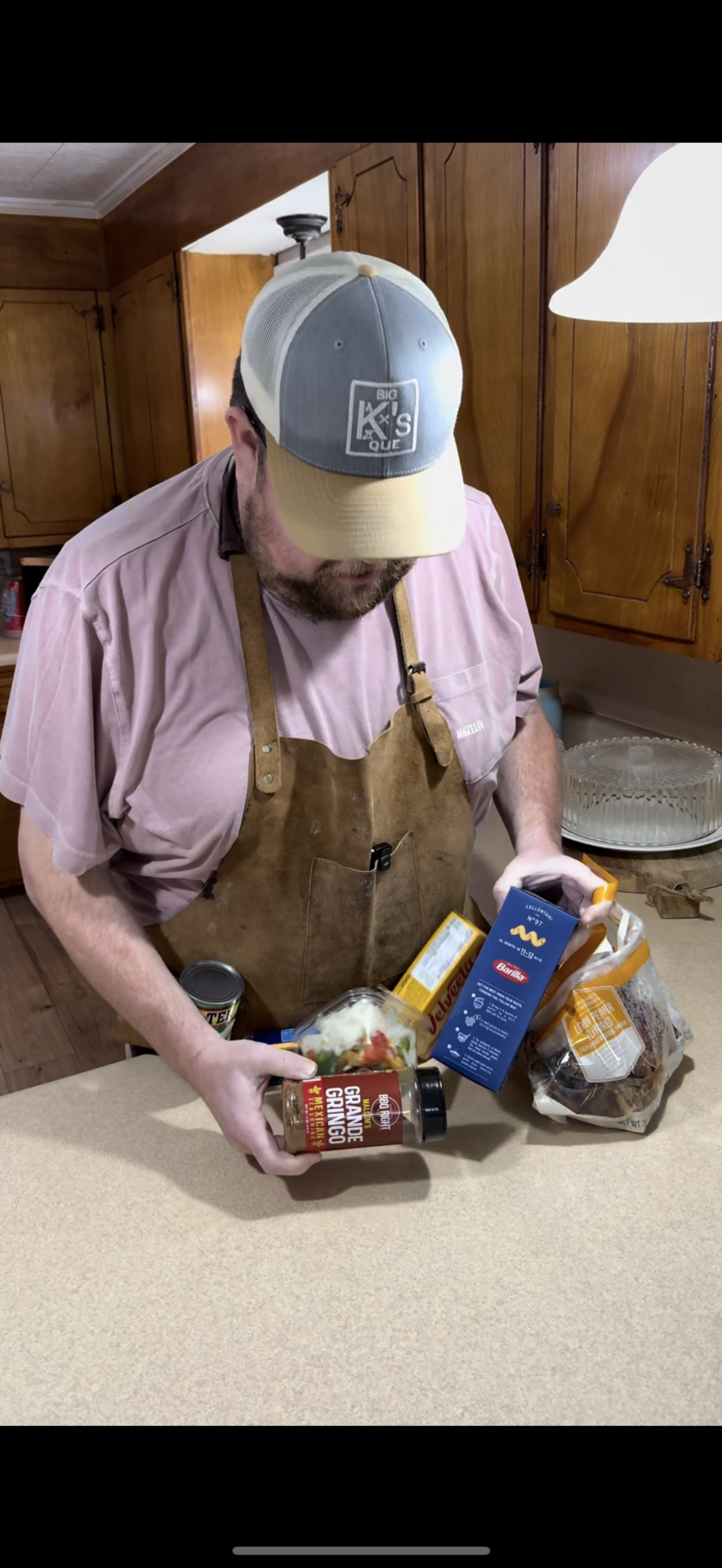 A man in a pink shirt, brown apron, and gray cap stands in a kitchen holding BBQ sauce, instant mashed potatoes, and canned food, with a grocery bag of items on the counter.