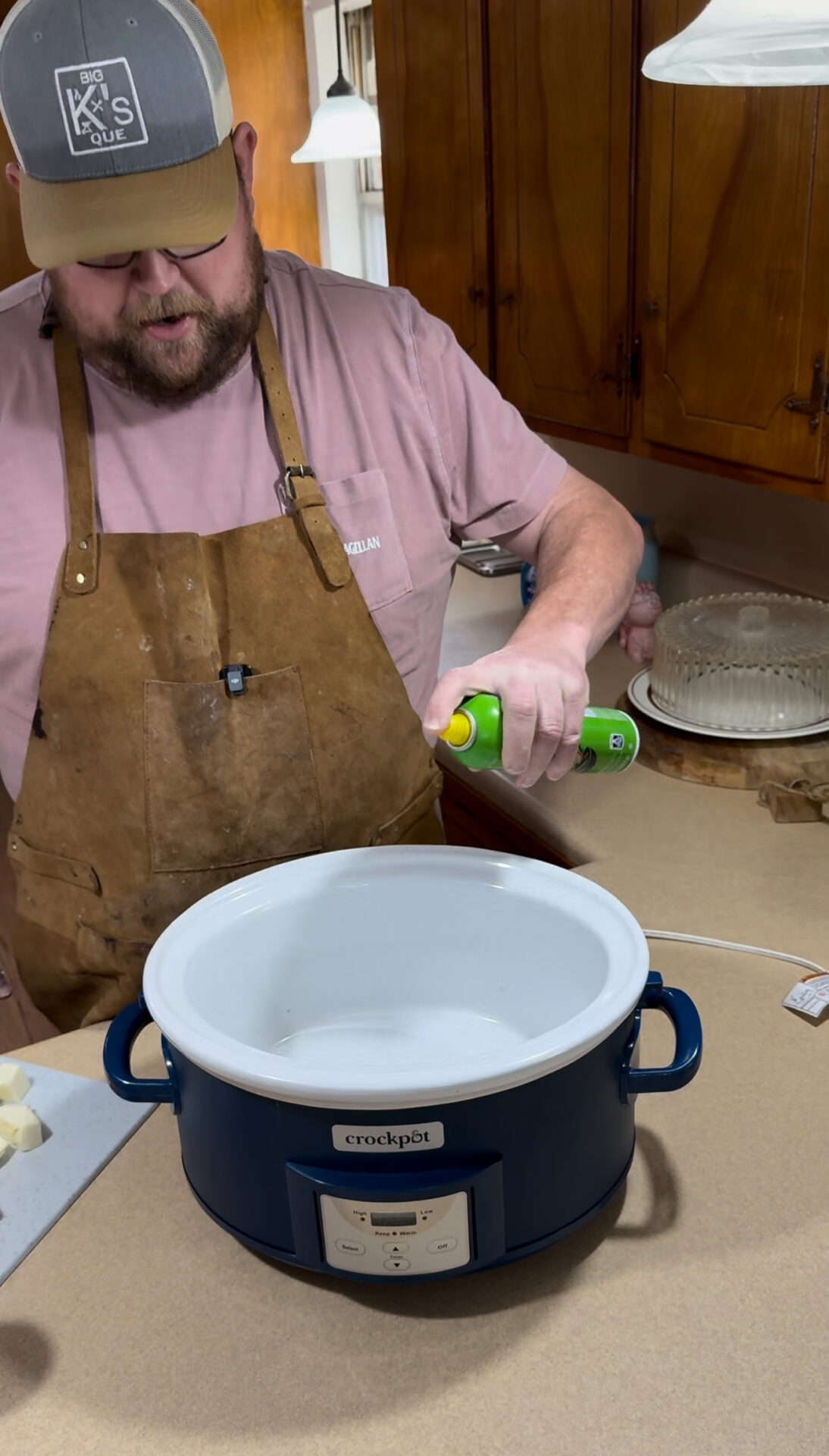 A man wearing a brown apron and a gray cap sprays cooking oil into a blue Crockpot slow cooker on a kitchen counter, with chopped onions and a cake in the background.