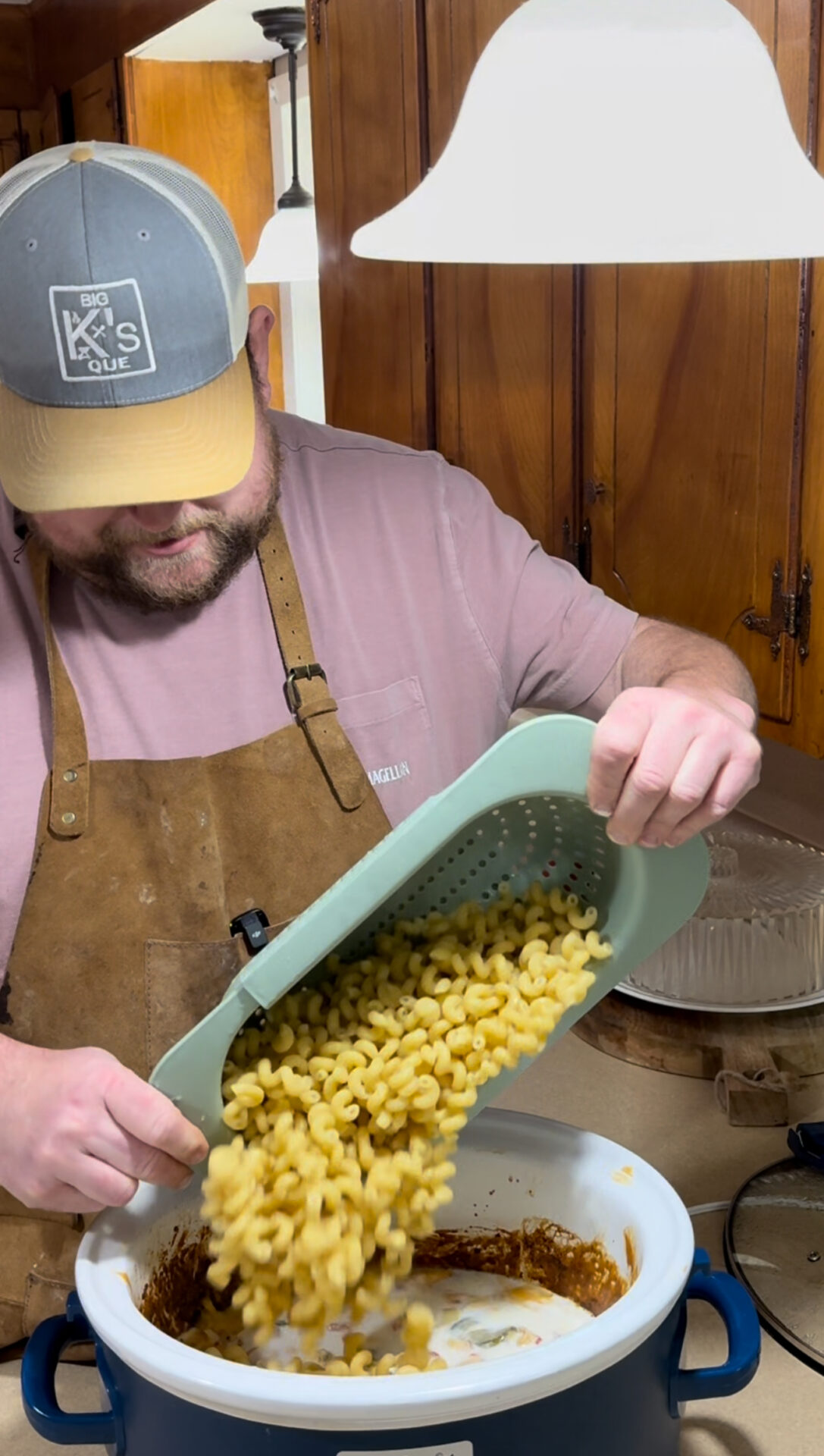 A man wearing a brown apron and a gray cap pours cooked pasta from a green colander into a white pot in a kitchen with wooden cabinets.