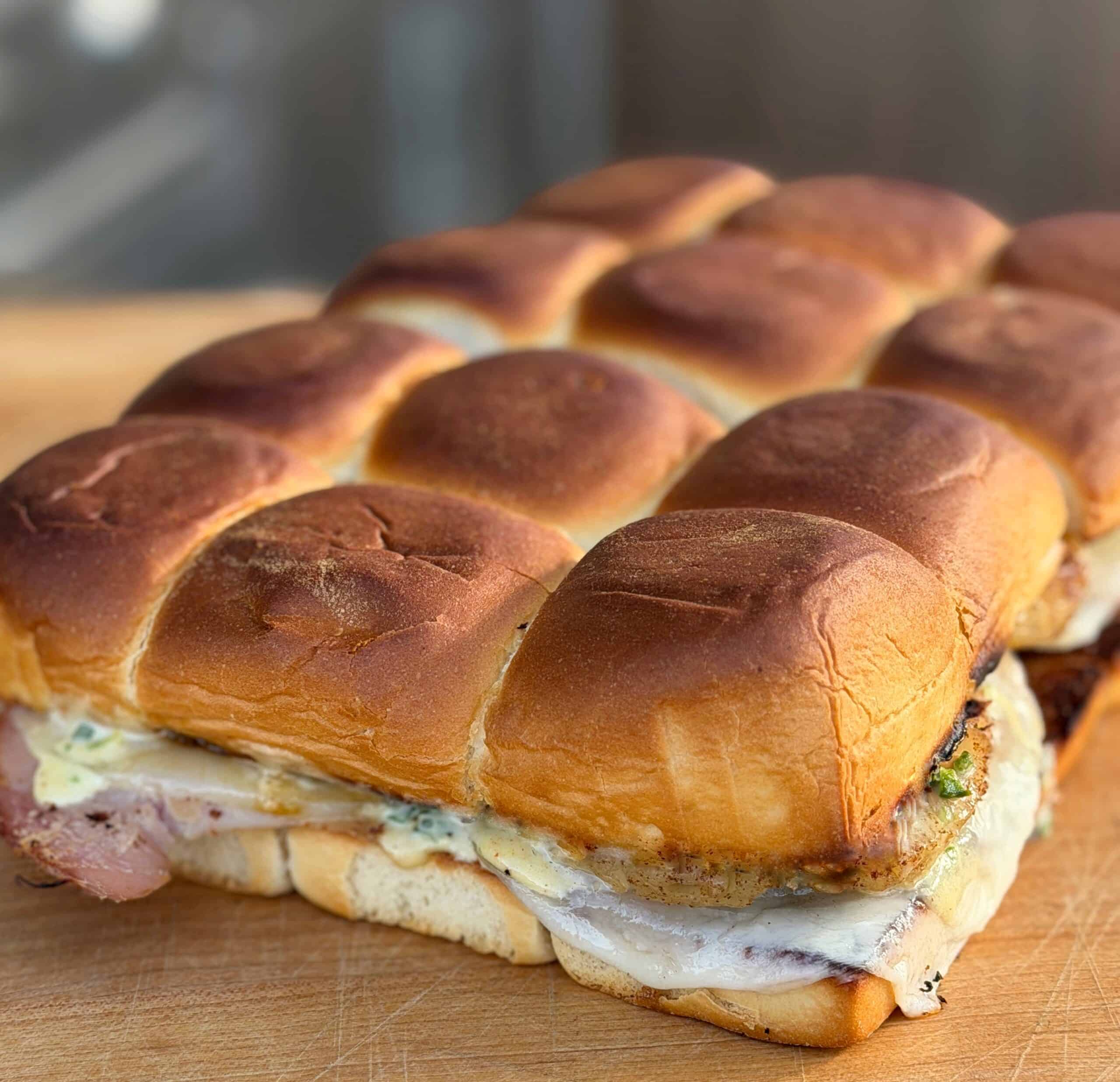 A close-up of a tray of golden-brown slider sandwiches filled with melted cheese, herbs, and meat, resting on a wooden surface.