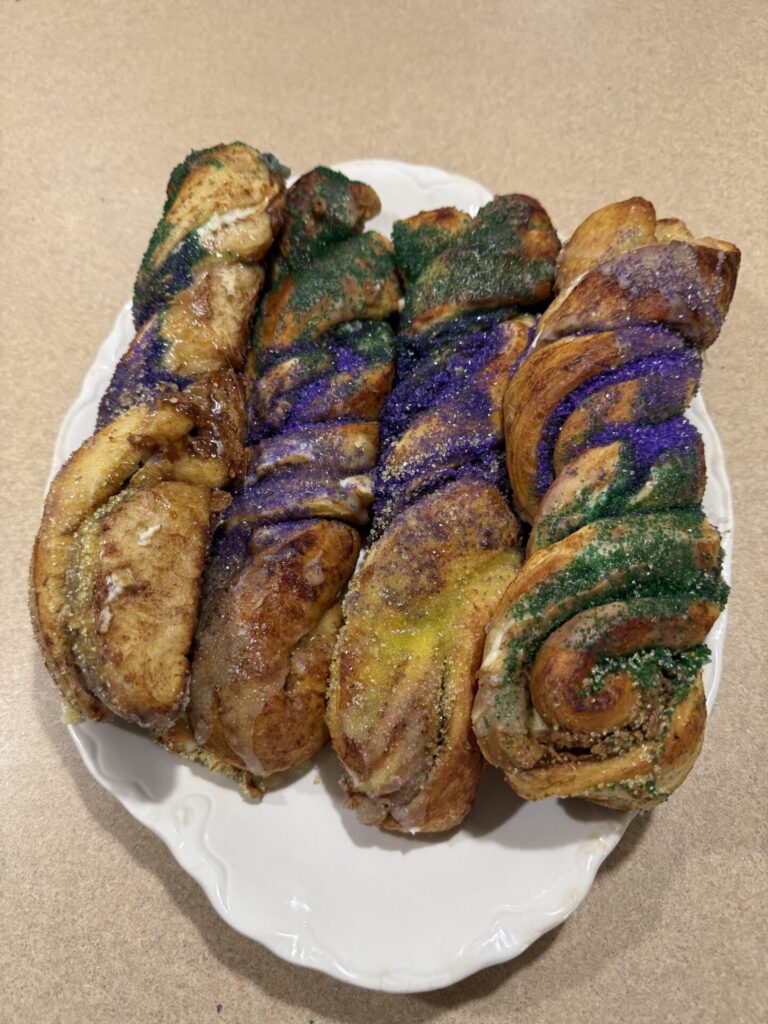 Four twisted pastries topped with green, purple, and yellow sugar are arranged on a white plate, set on a light-colored countertop. The pastries appear festive and colorful.