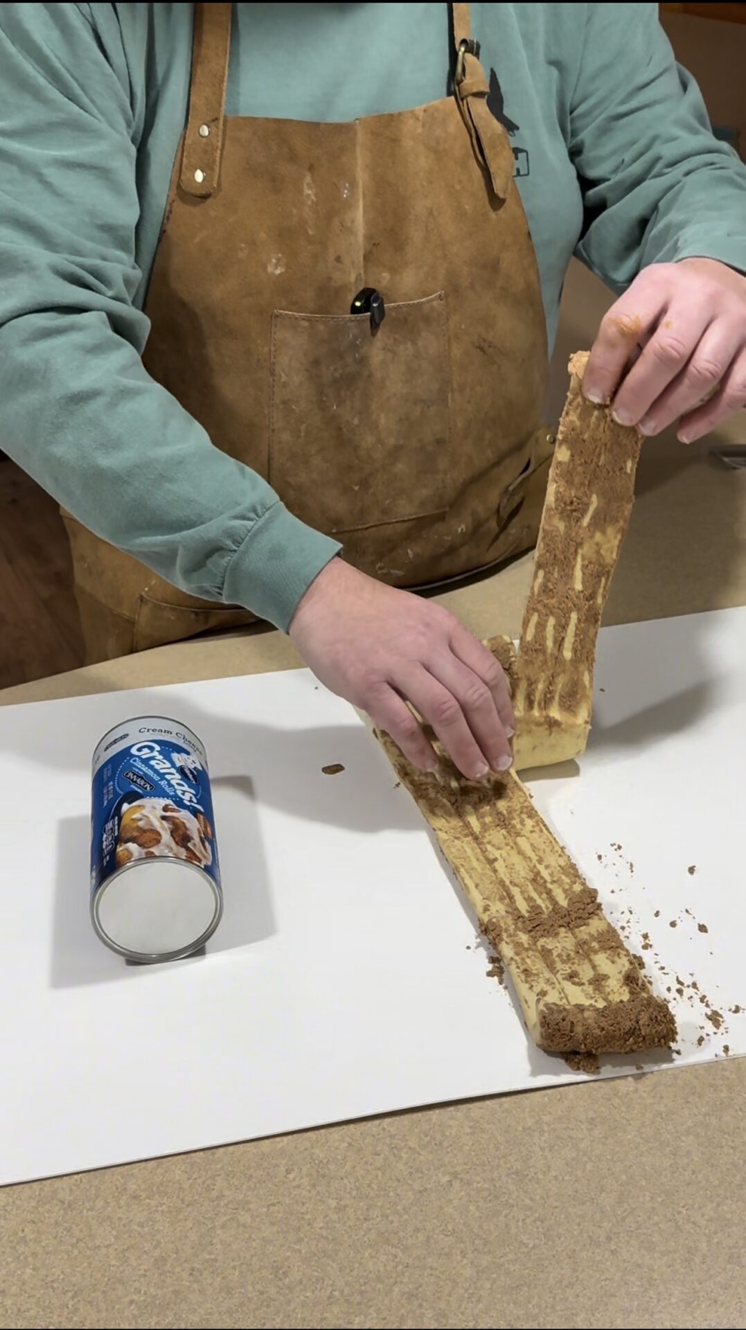 A person in a brown apron unrolls cinnamon roll dough onto a white surface, with a can of store-bought cinnamon rolls placed nearby.