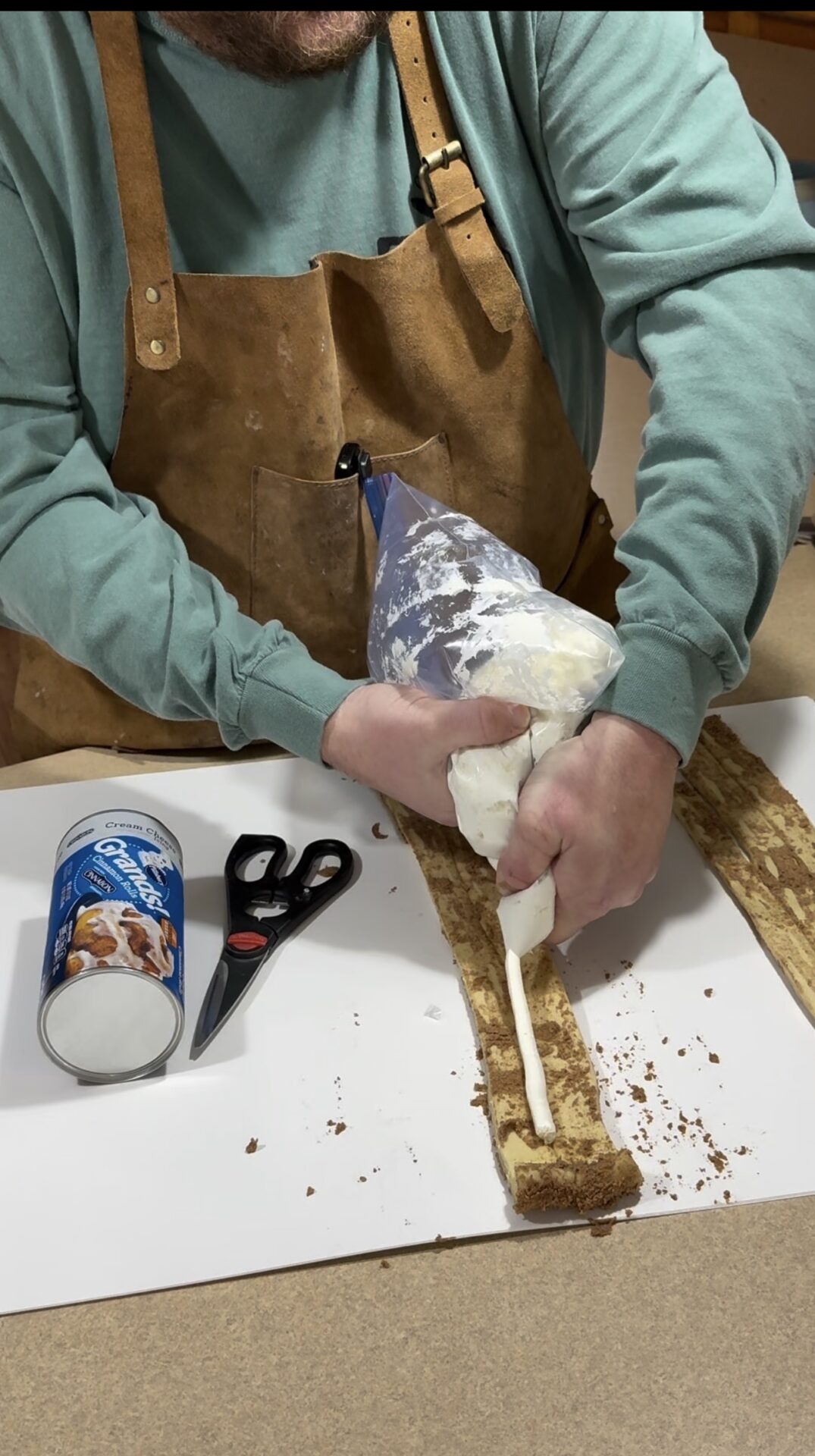 A person in a brown apron pipes white filling onto a strip of cinnamon roll dough. An open can of cinnamon rolls and scissors are on the table next to them.