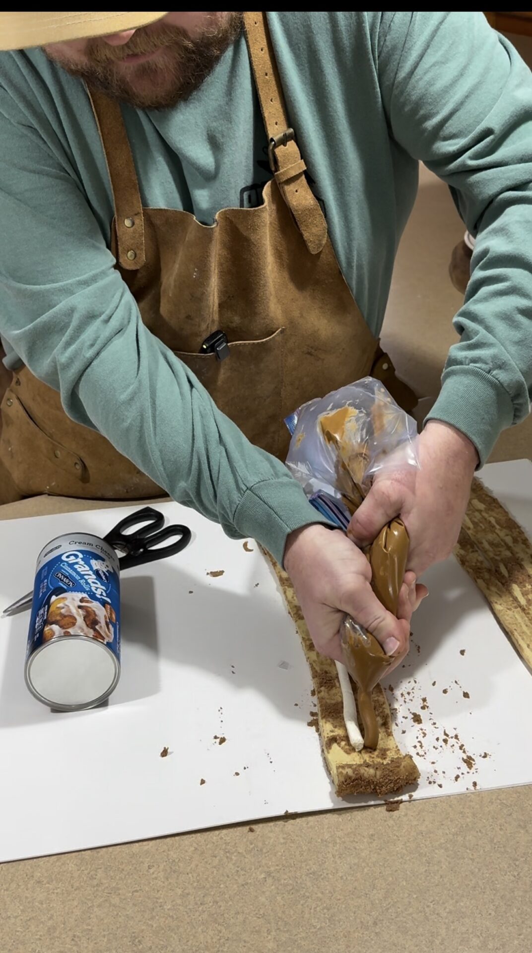 A person wearing a brown apron and green shirt pipes caramel filling onto a strip of baked dough on a counter. An open can of cinnamon rolls and a pair of scissors are next to the dough.