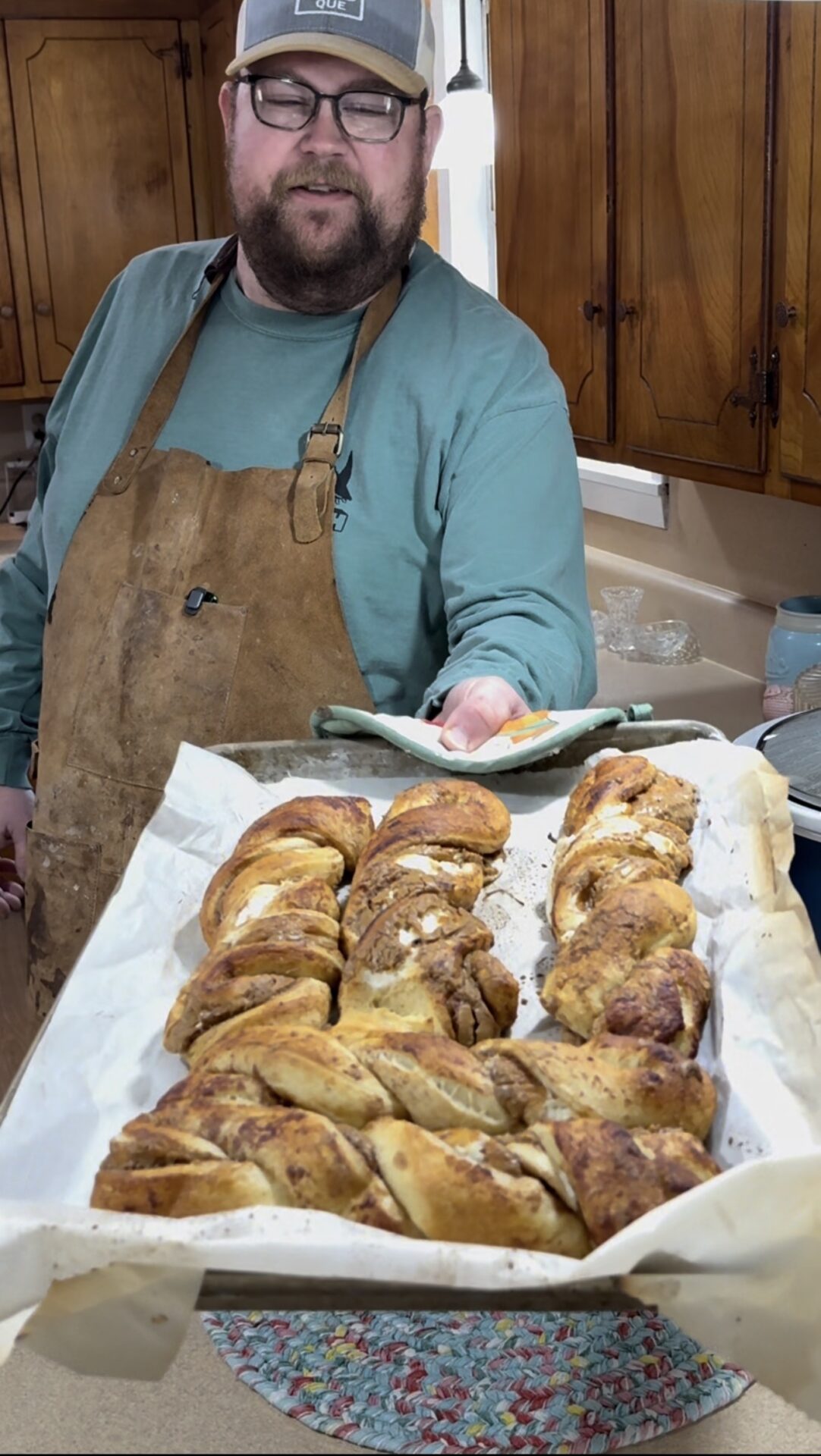 A man wearing glasses, a hat, and an apron holds a tray of freshly baked, twisted bread on parchment paper in a kitchen with wooden cabinets.
