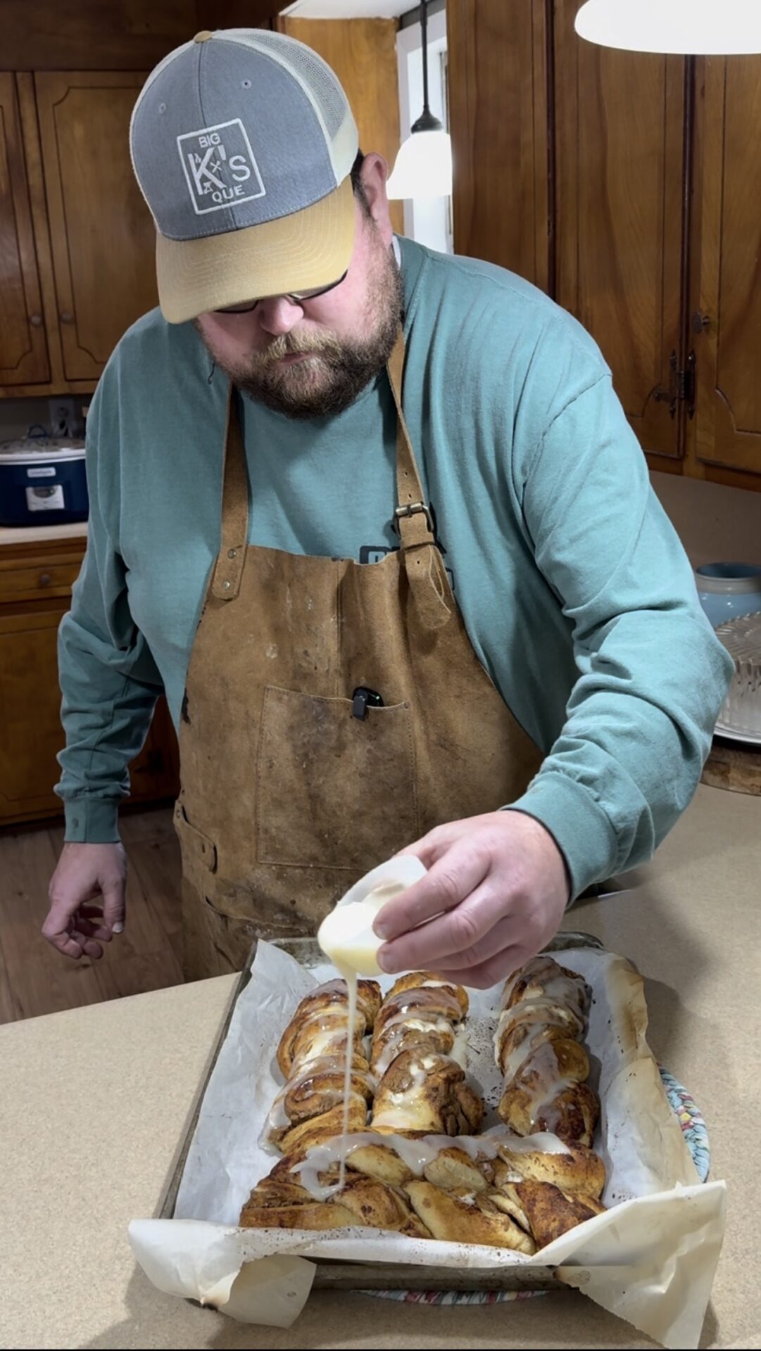 A man wearing a blue shirt, brown apron, and gray cap pours icing over freshly baked cinnamon rolls on a parchment-lined baking sheet in a kitchen.