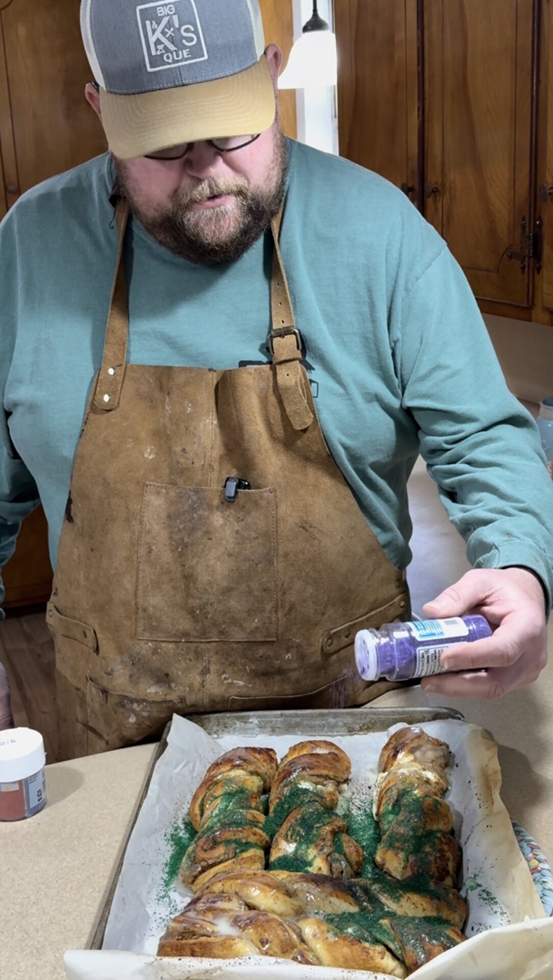 A bearded man wearing glasses, a brown apron, and a blue-green shirt sprinkles purple sugar on a baked King Cake topped with green sugar and icing in a home kitchen.
