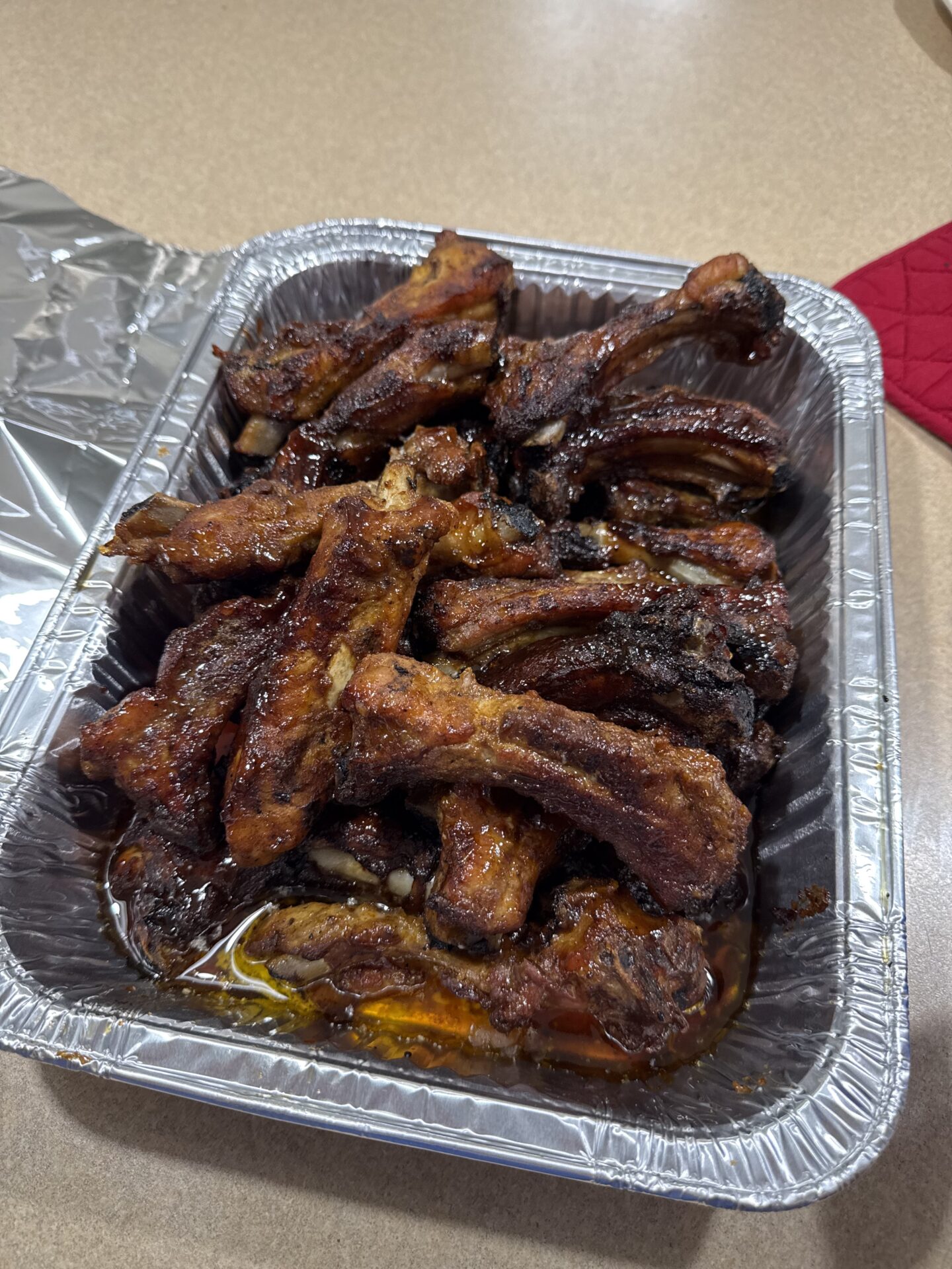 A disposable aluminum tray filled with cooked, saucy pork ribs sits on a countertop next to a piece of crumpled aluminum foil and a red oven mitt. The ribs are glazed and slightly charred.