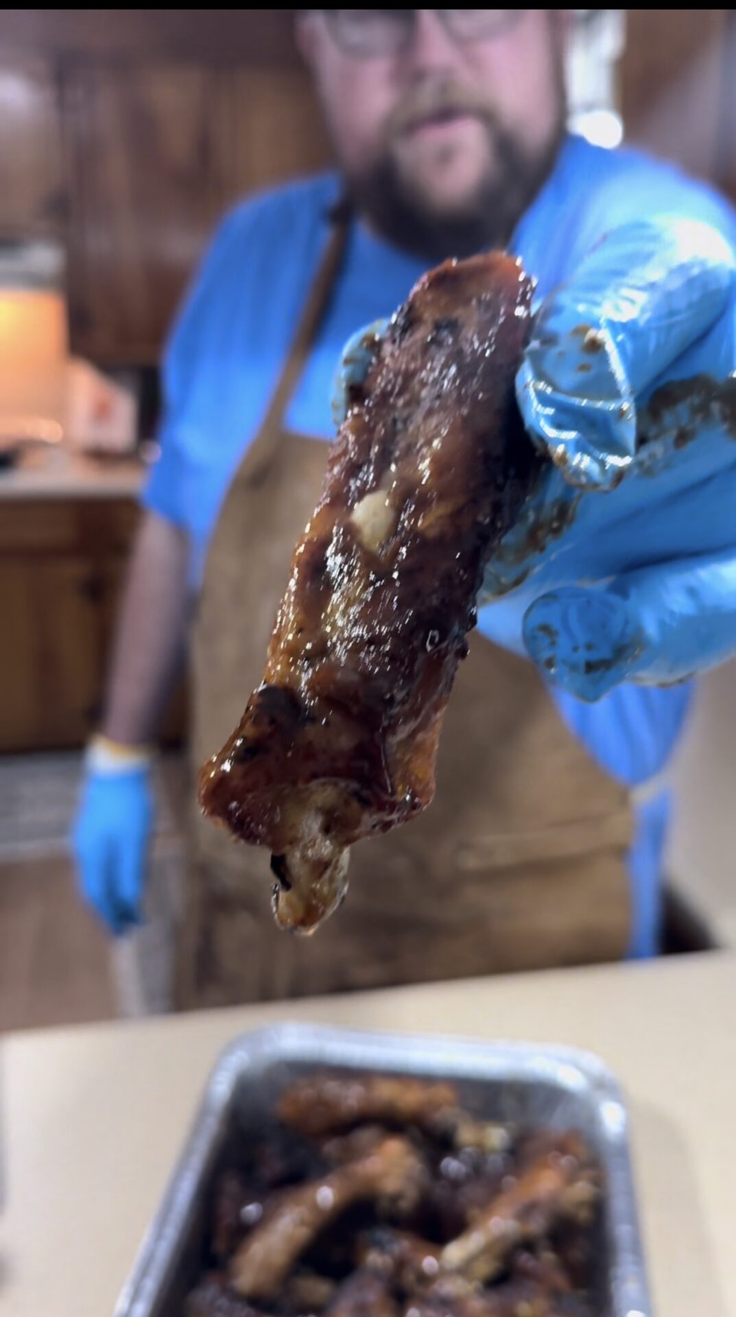 A person wearing a blue glove and brown apron holds up a saucy piece of cooked meat, possibly a rib or chicken wing, over a tray of similar pieces in a kitchen. The background is slightly out of focus.
