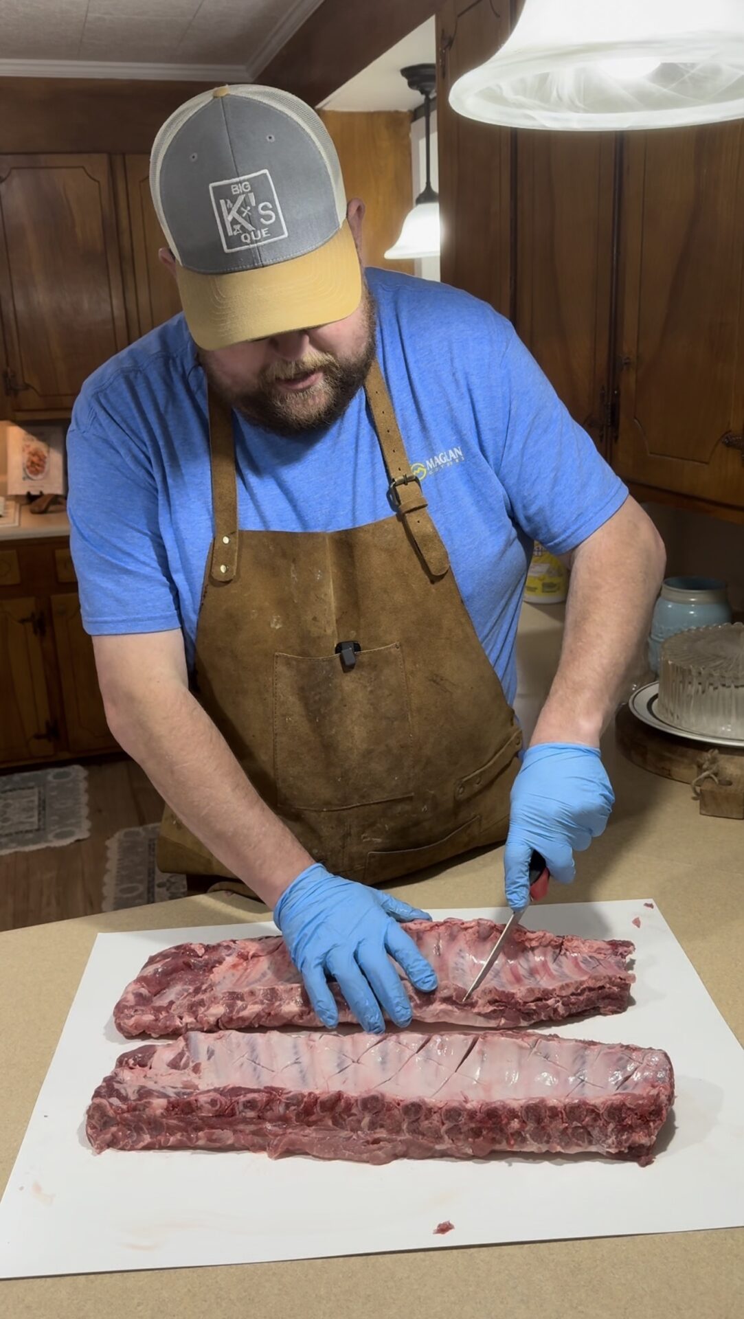 A man wearing a brown apron, blue gloves, and a gray cap slices raw pork ribs on a white cutting board in a kitchen with wooden cabinets.