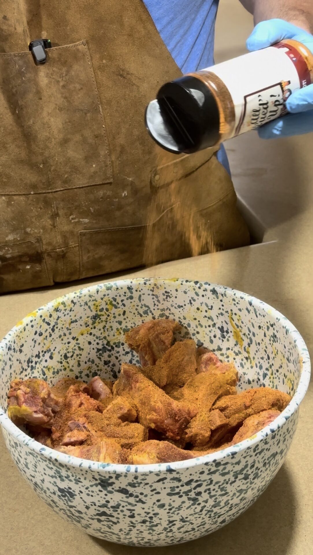 A person in an apron sprinkles seasoning from a bottle onto raw chicken wings in a speckled bowl, preparing them for cooking.