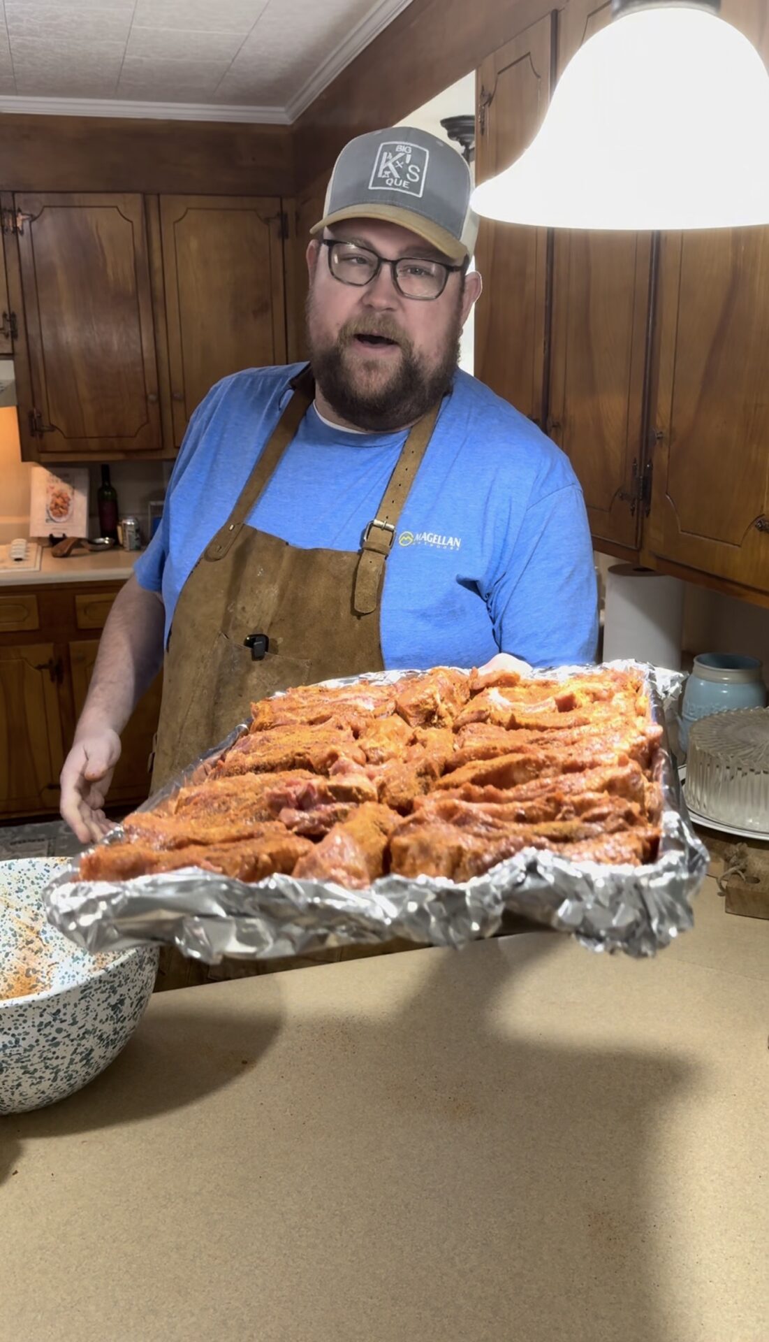 A man wearing glasses, a blue shirt, brown apron, and a gray cap holds a large tray of seasoned meat covered with foil in a kitchen, standing next to a mixing bowl and a counter.